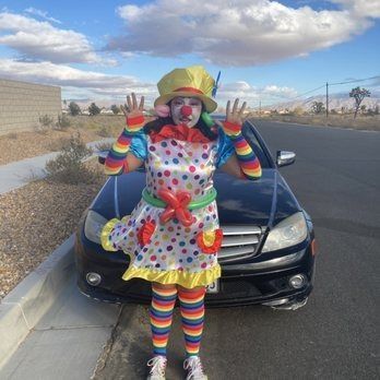 Clown in a polka-dot dress raises hands by a black car on a sunny day.