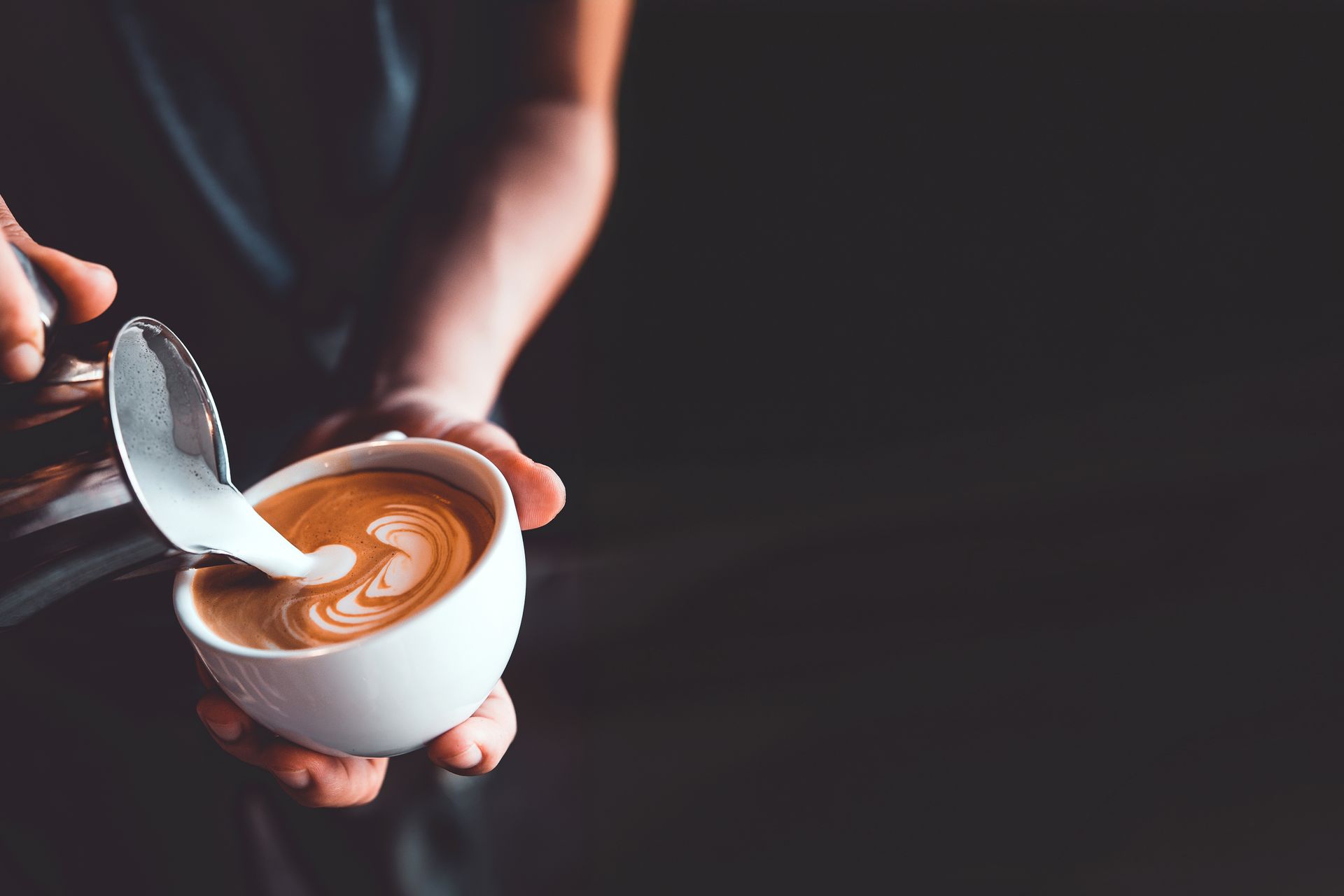 Hands holding a white cup of coffee with latte art and a spoon over a dark background