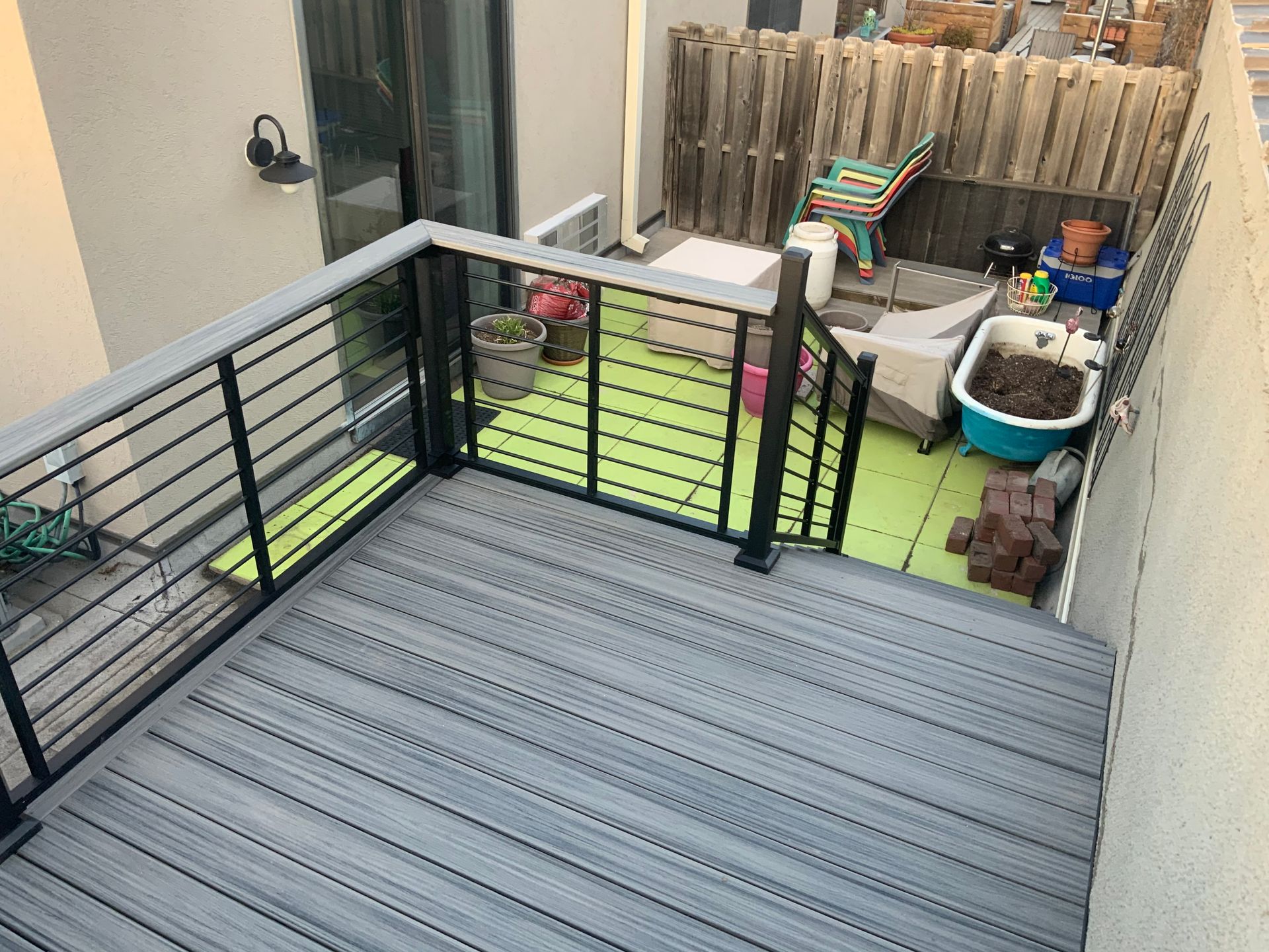 Backyard deck with gray flooring, black railing, and a small patio area with furniture and potted plants.