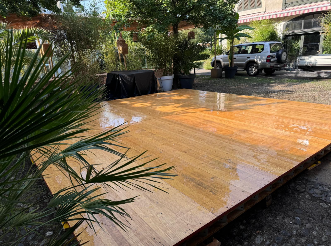 Terrasse en bois dans un jardin, partiellement inondée. Plantes vertes et voiture argentée visibles.