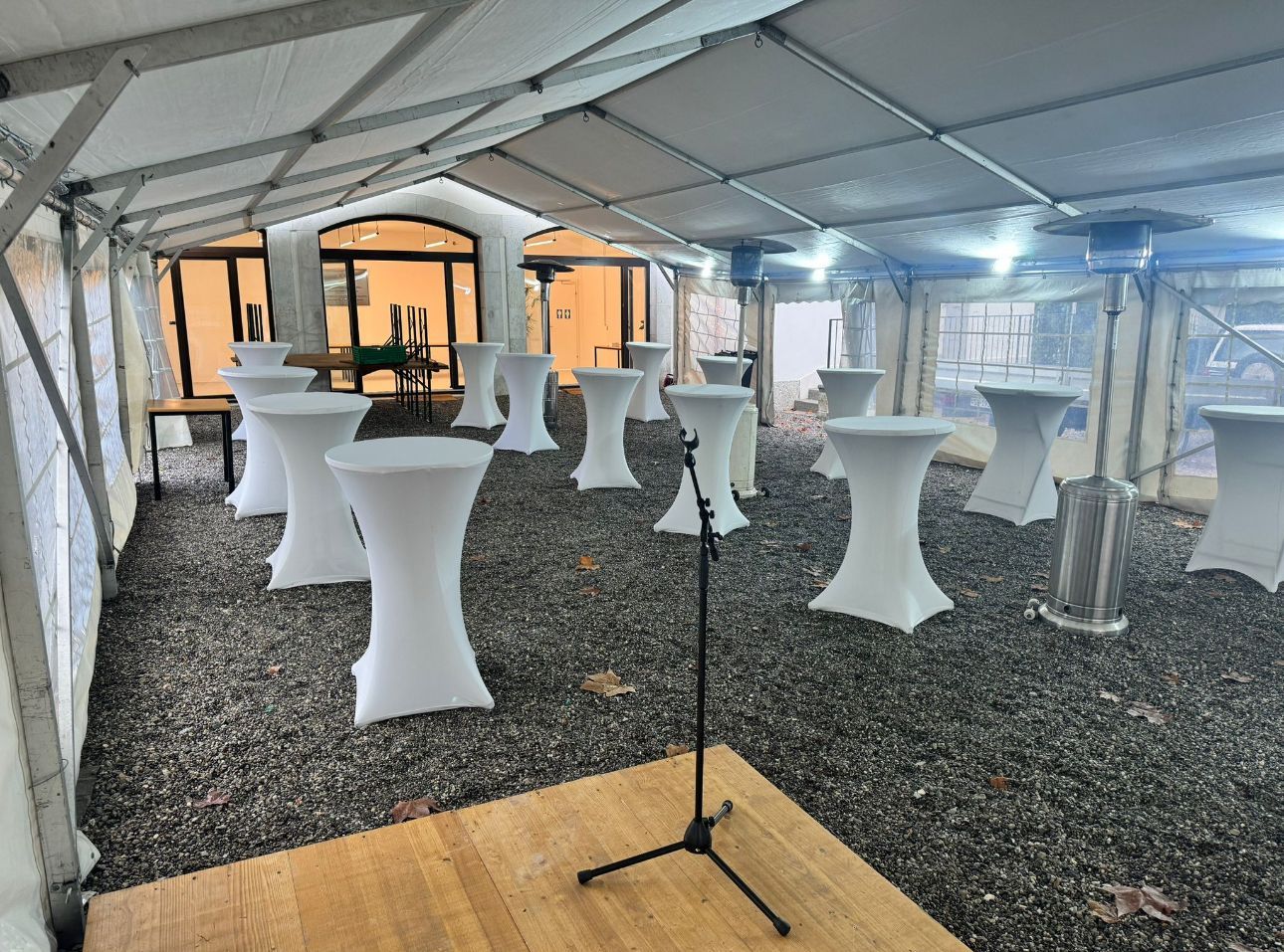 Sous une tente, des tables basses blanches sont disposées sur un sol gravillonné. Un pied de micro est installé sur une estrade en bois.