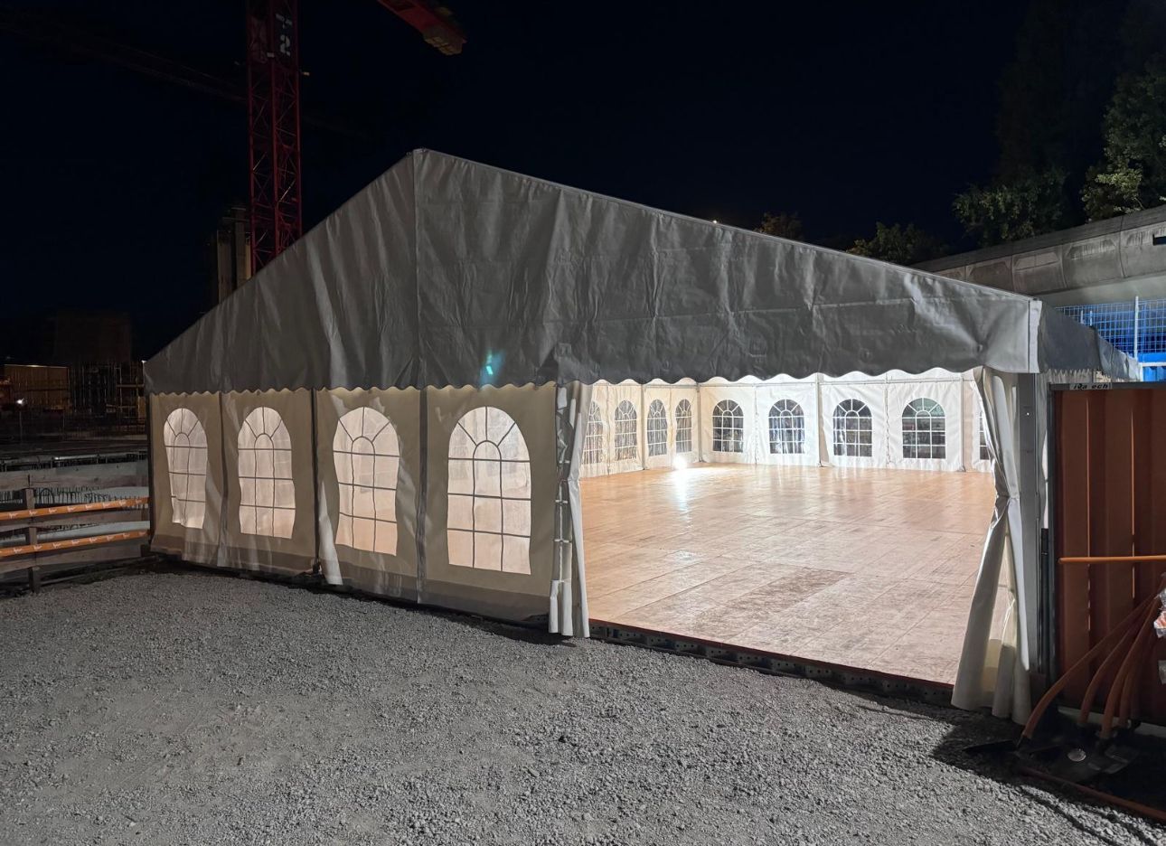 Grande tente blanche installée sur du gravier, éclairée à l'intérieur. Chantier de construction en arrière-plan.