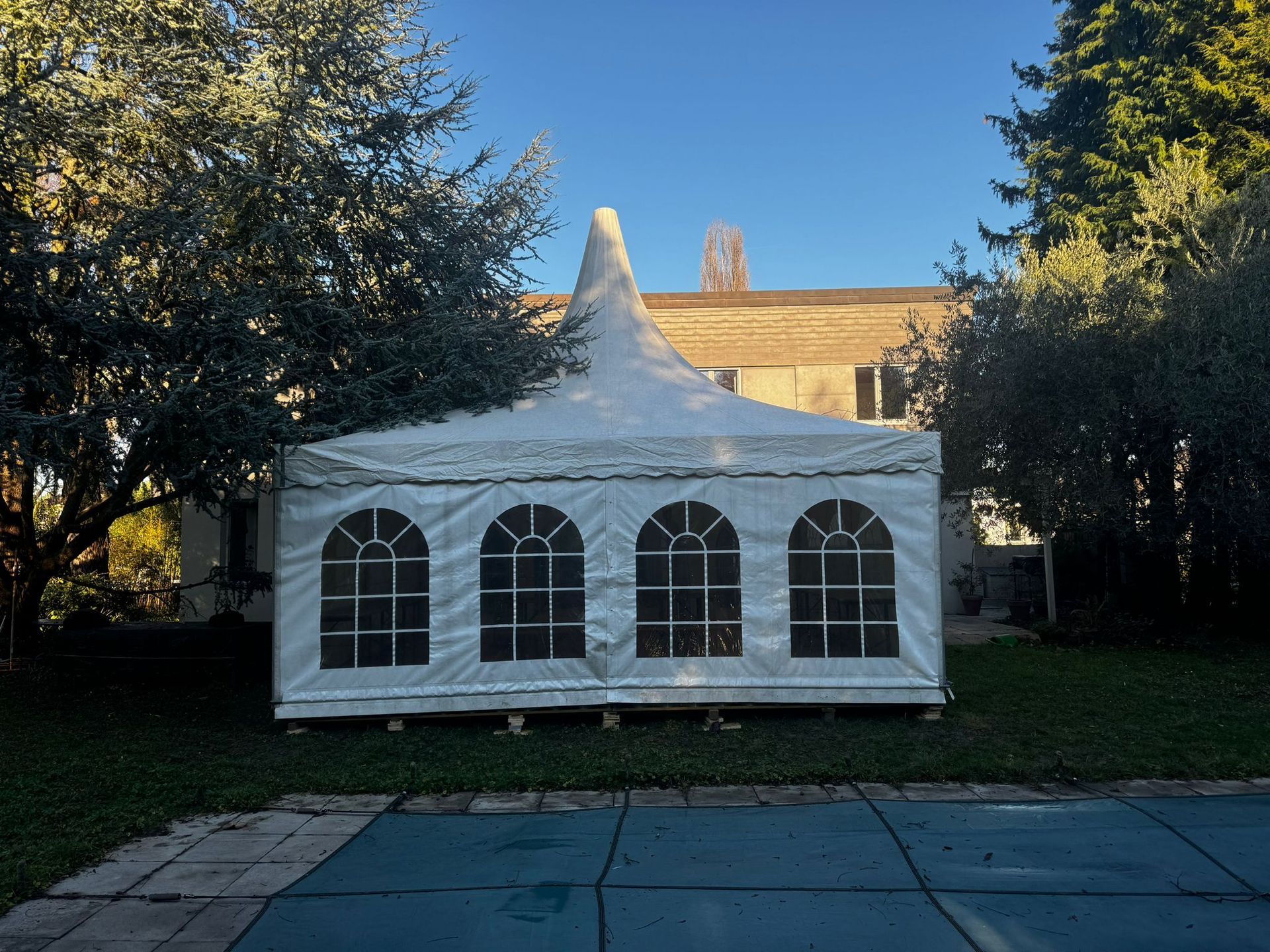 Tente événementielle blanche avec fenêtres cintrées, installée sur l'herbe près d'une piscine, avec un bâtiment en arrière-plan.