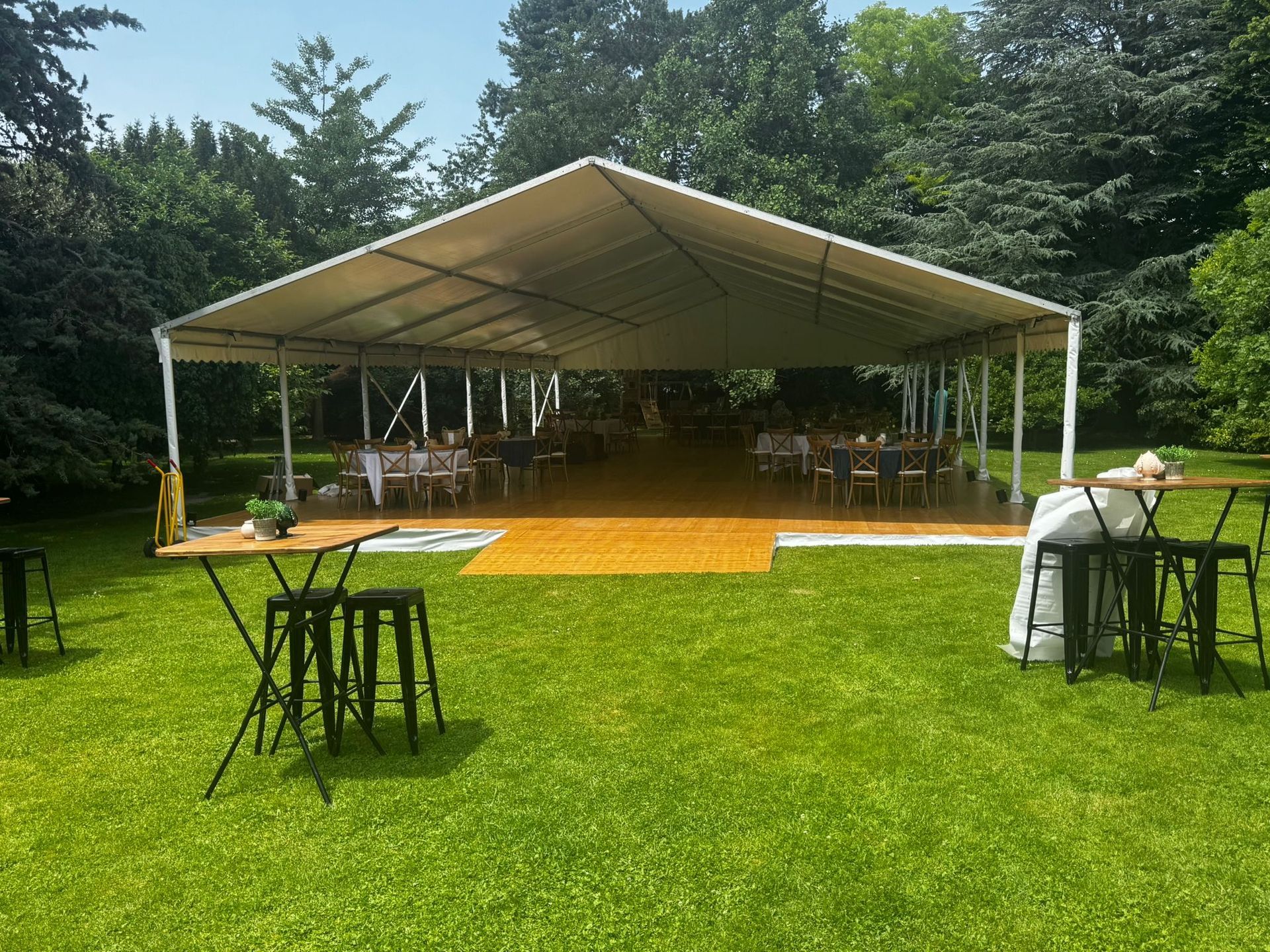 Une tente est installée pour un événement dans un espace extérieur herbeux, avec des tables, des chaises et des arbres en arrière-plan.