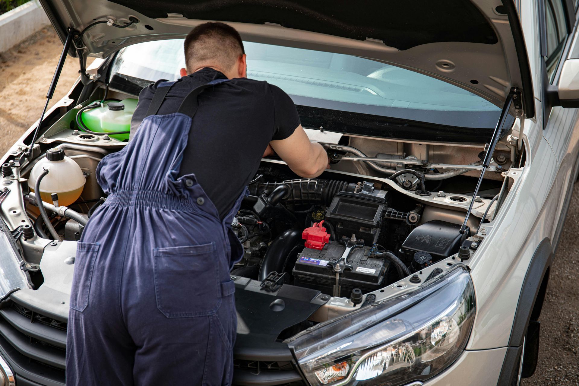 A person in blue coveralls and a black t-shirt leaning over an open car hood to perform engine maintenance.