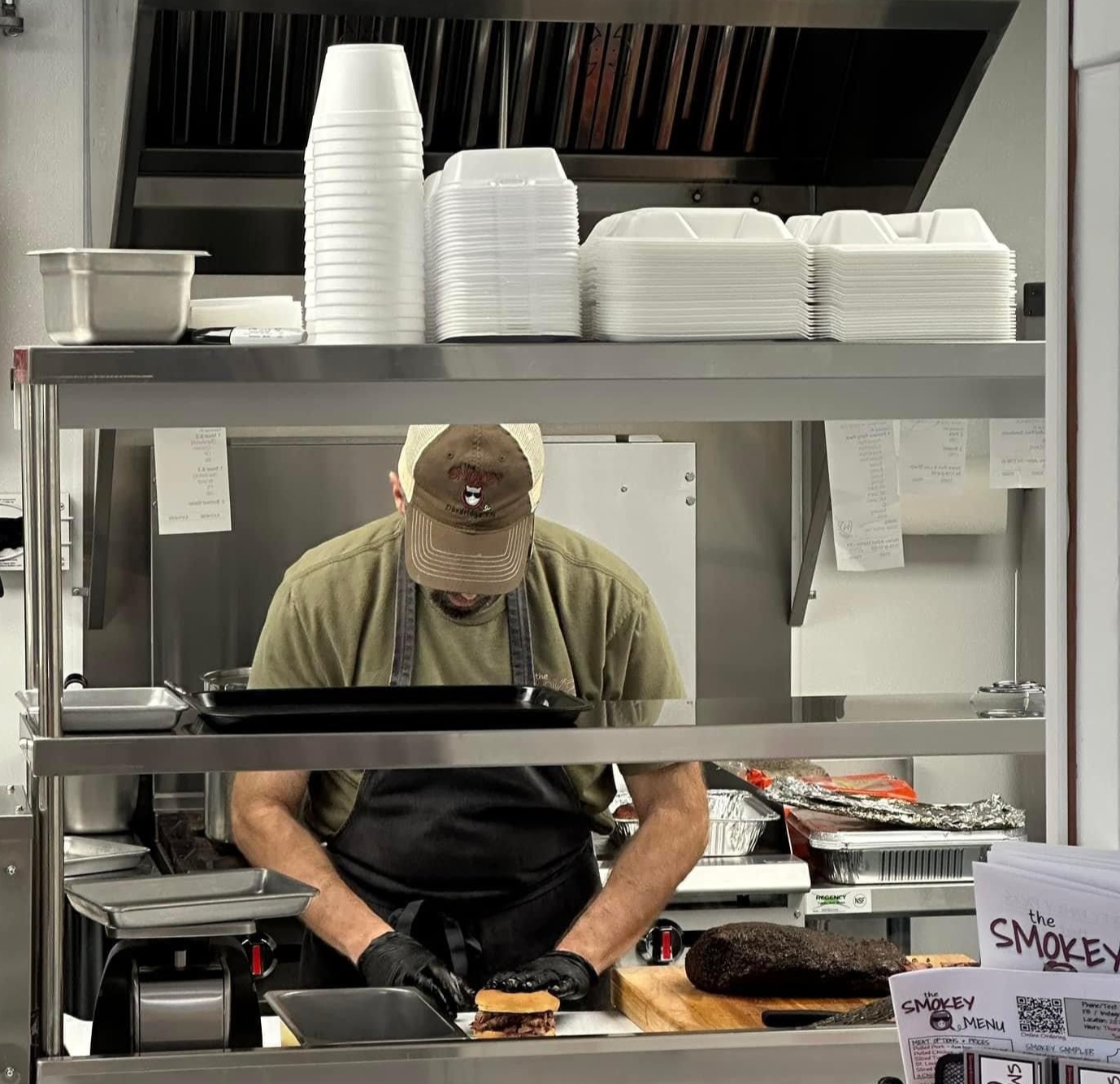 A styrofoam container filled with meat , beans , macaroni and cheese , and pickles.