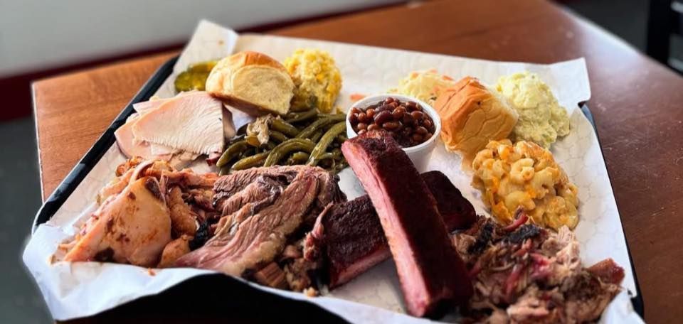 A styrofoam container filled with meat , beans , macaroni and cheese , and pickles.
