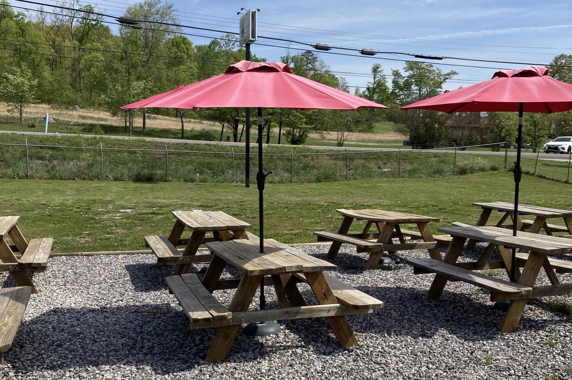 Outdoor picnic tables with red umbrellas on gravel, near a green space and road.