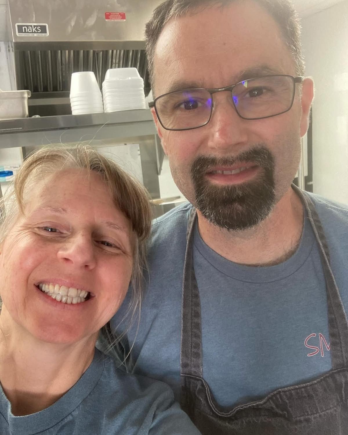 Smiling man and woman in a kitchen; man has glasses and beard.