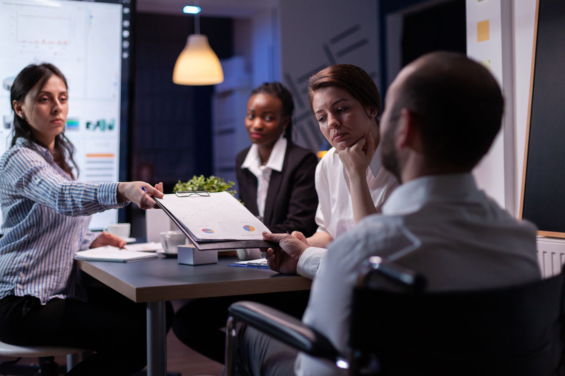 People in a meeting room, a woman hands paperwork to a man in a wheelchair, other colleagues watch.