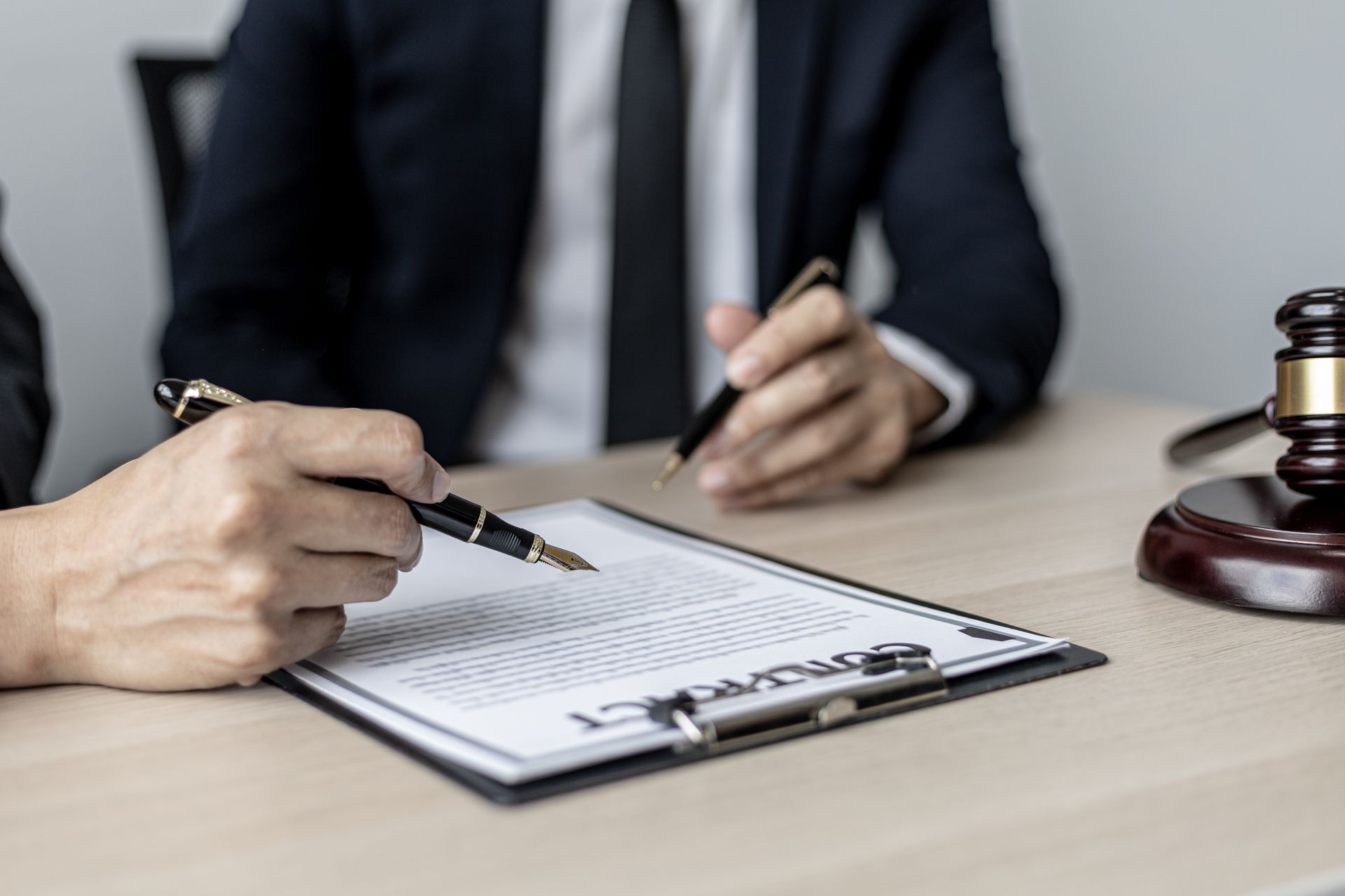 Two people in suits at a table; one points at a document. A gavel and tablet sit nearby.