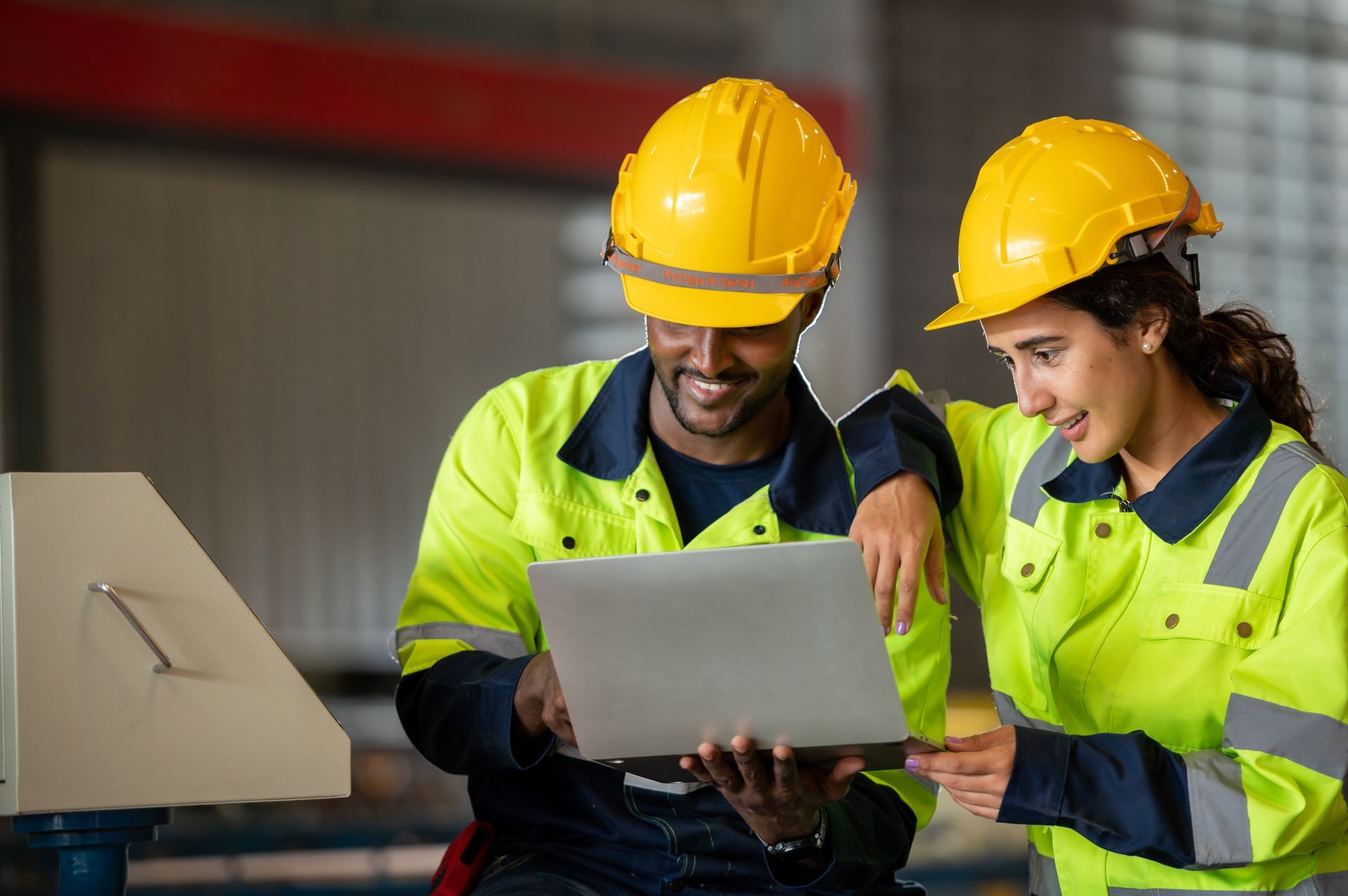 Four people in safety vests and hard hats look at a laptop and tablet in a warehouse setting.