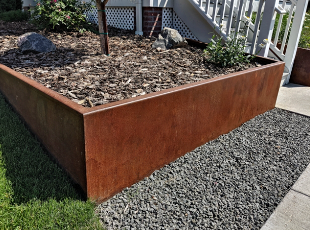 Rusty metal raised garden bed with plants, mulch, and gravel border next to a sidewalk.
