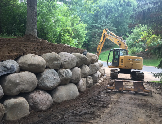 Yellow excavator next to a stacked stone retaining wall. Trees in background, dirt path in front.