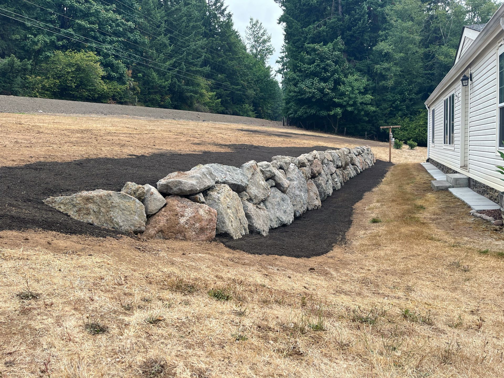 A long stone wall, surrounded by brown mulch and dry grass, separates the yard from a house.