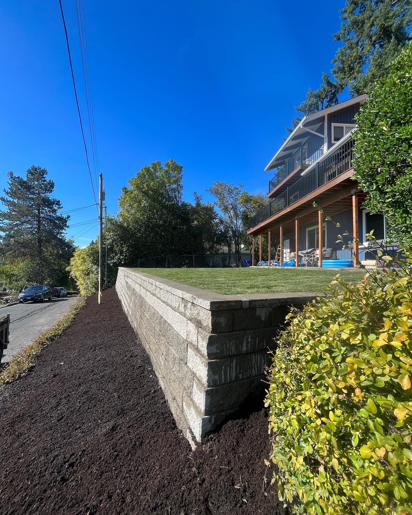 Stone retaining wall with grass top, mulch, and a house in the background. Blue sky and trees.