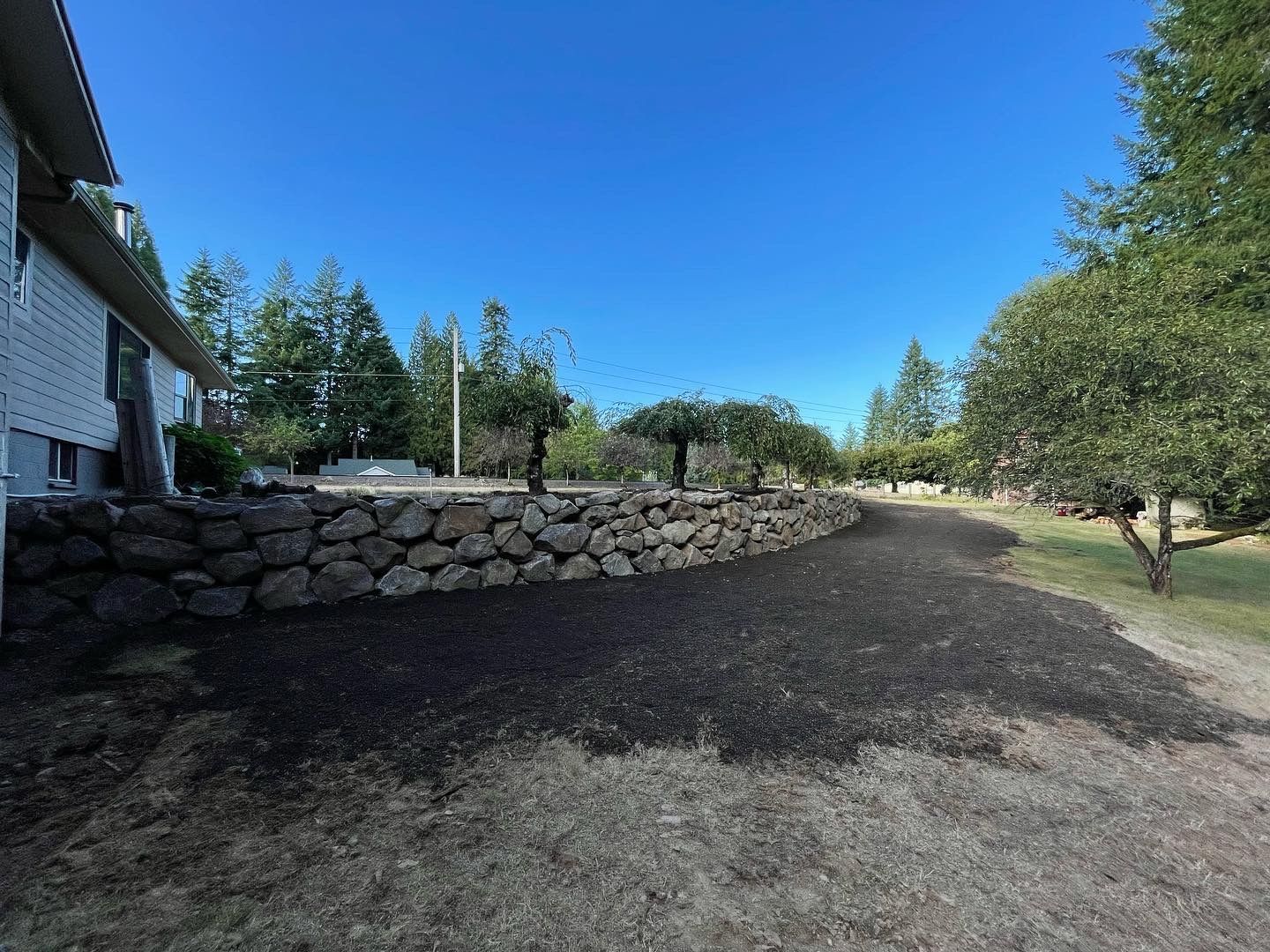 A stone retaining wall in front of a house, dark soil, trees, and blue sky.