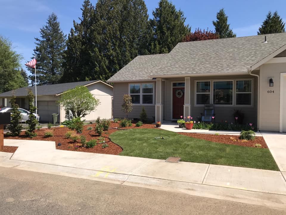 A light tan house with red door and green lawn, brown mulch, and an American flag.