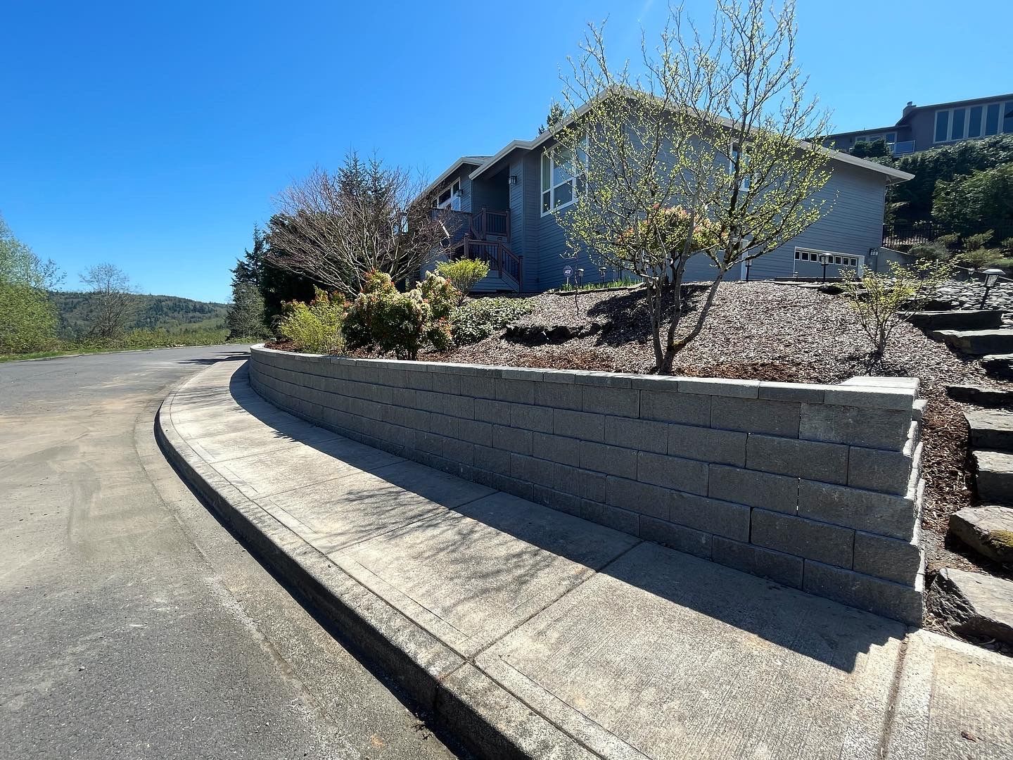Gray block retaining wall next to a road and sidewalk with a house in the background.