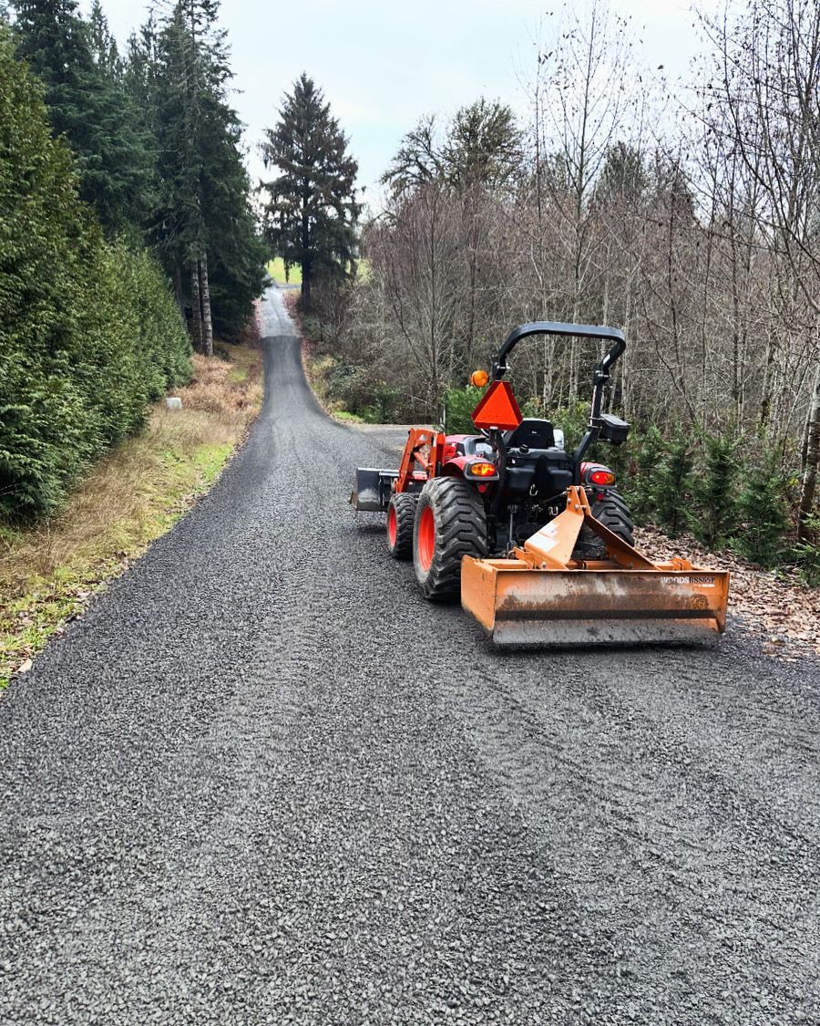 A tractor grading a gravel road on a tree-lined hillside. Orange tractor, gray gravel, green trees.