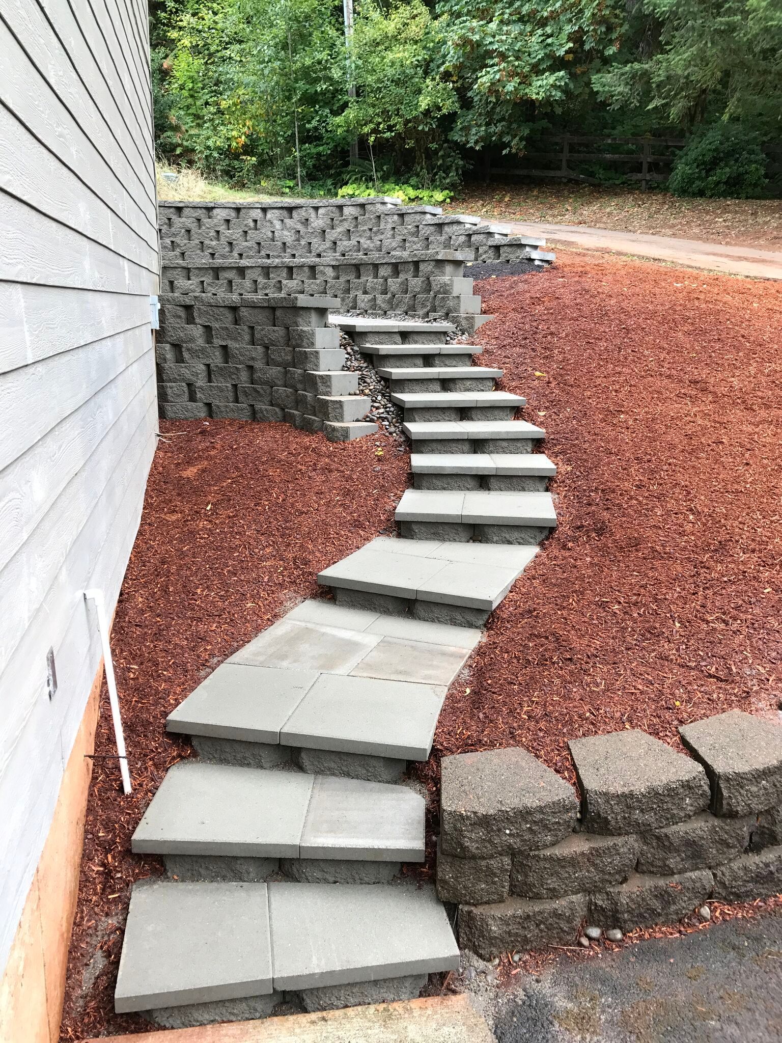 Stone steps wind up a hillside, bordered by a block retaining wall and reddish-brown mulch.