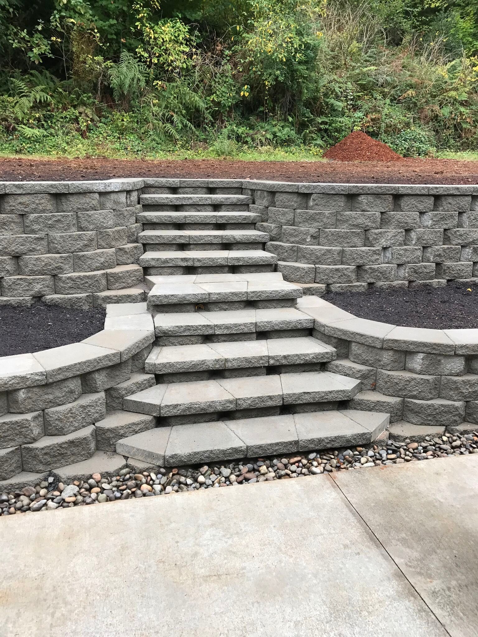 Stone steps and retaining wall leading up to a wooded area, with black mulch and gravel below.