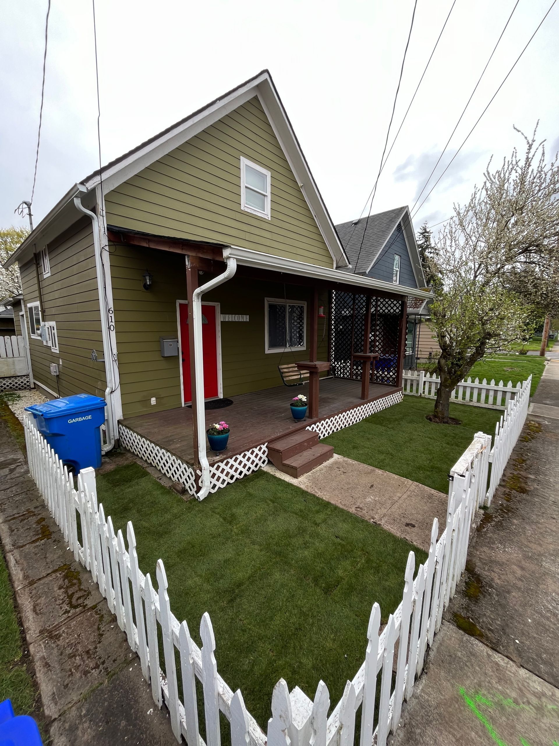 Green house with white picket fence, red door, and porch. Blue trash bin in front yard.