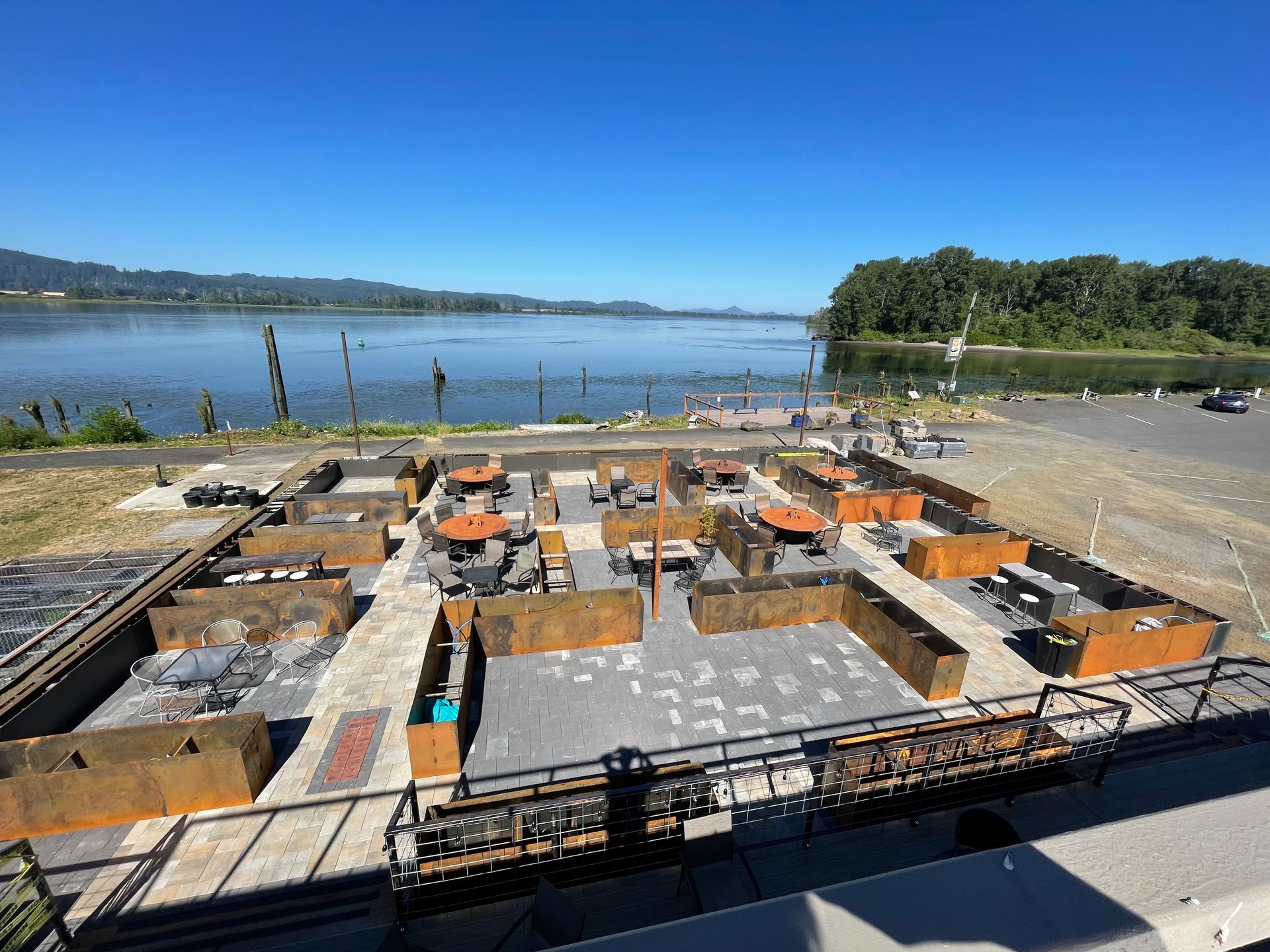 Outdoor restaurant patio with tables, seating, and metal dividers overlooking a river on a sunny day.
