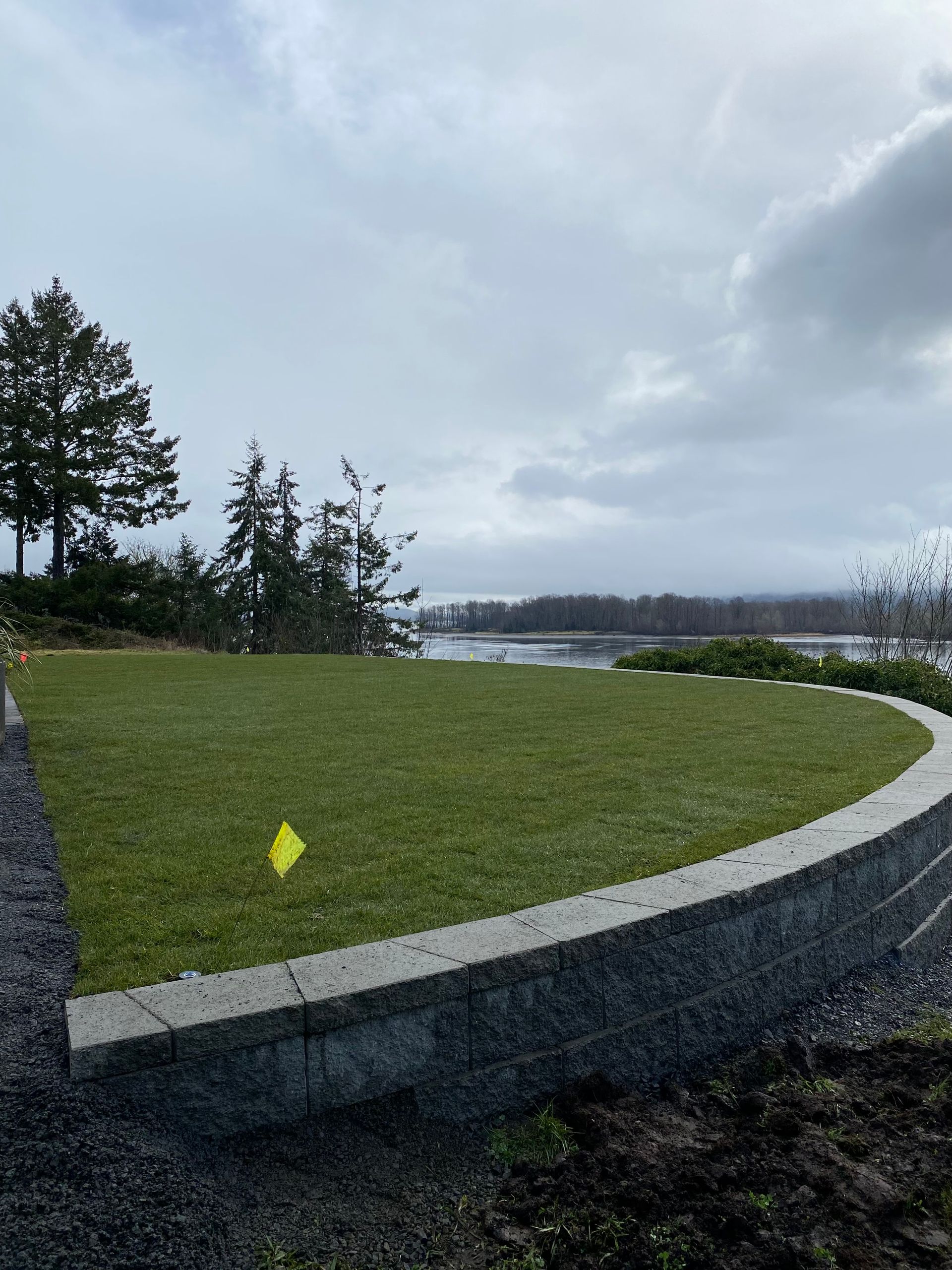 Lush green lawn bordered by a stone wall overlooks a body of water under a cloudy sky.