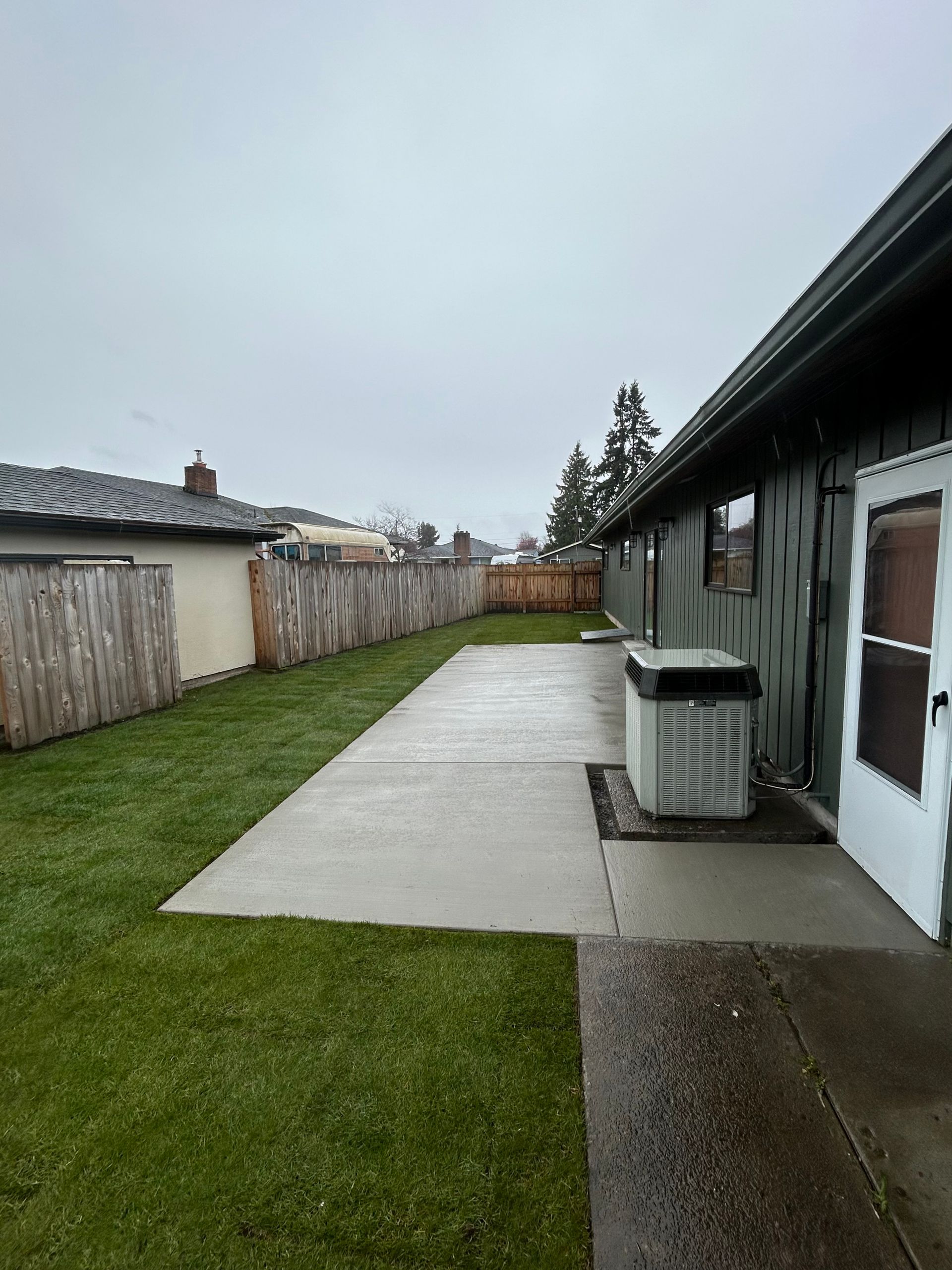 Backyard with green lawn, concrete patio, wooden fence, and a house. Overcast day.