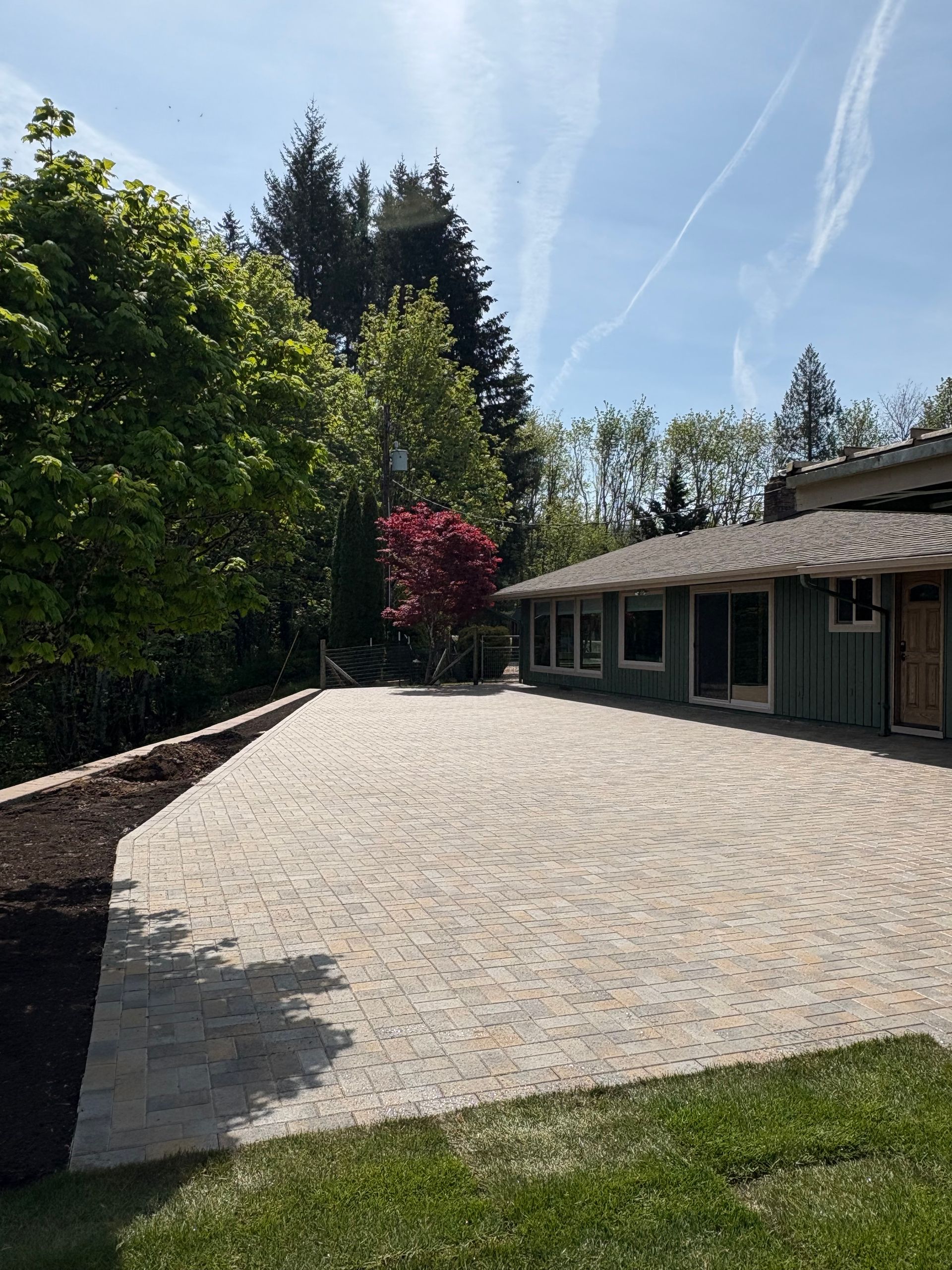 Brick patio in front of a green house, surrounded by trees, under a blue sky.