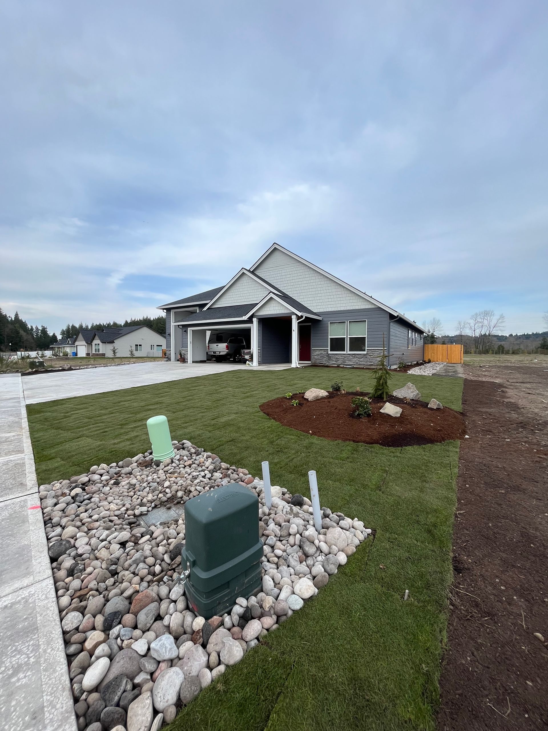 Gray house with attached carport, green lawn, gravel, and cloudy sky.