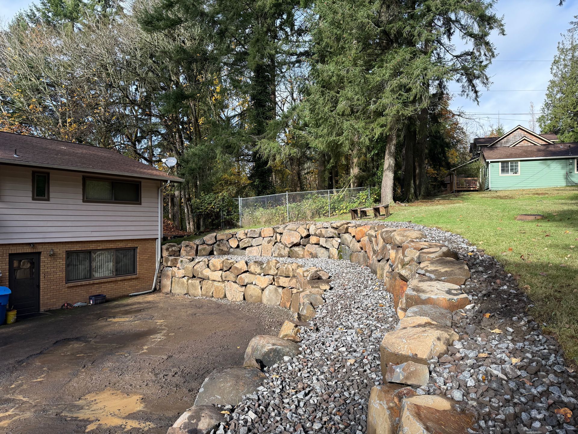 A stone retaining wall in a yard, gravel path, house on left, green house in the distance.