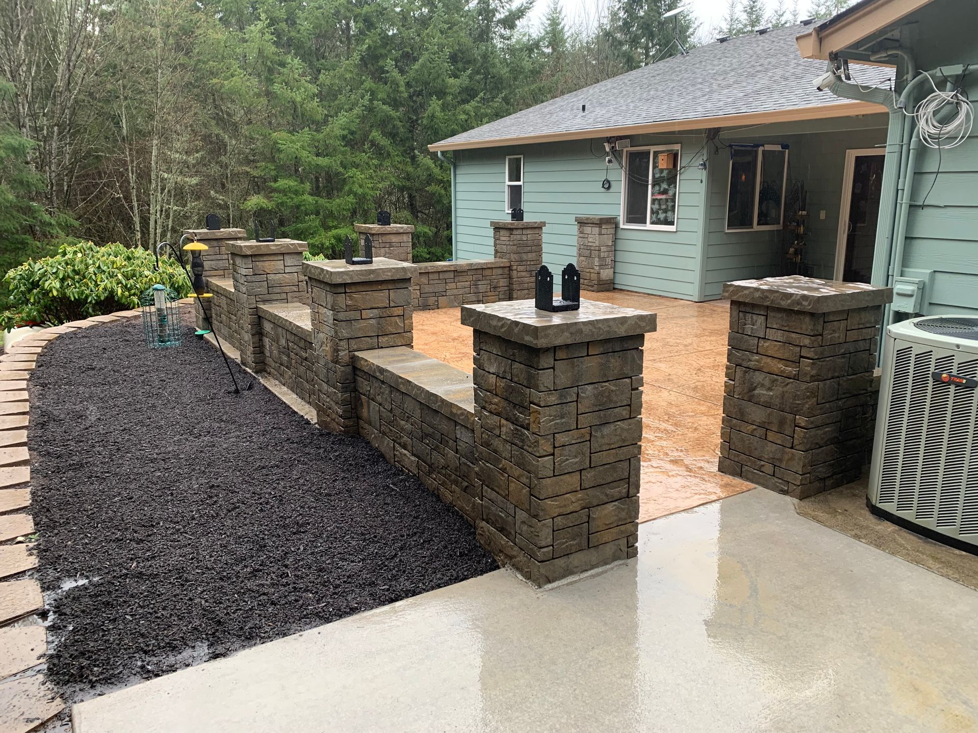 A stone wall and patio surround a house with a dark mulch bed in the foreground.