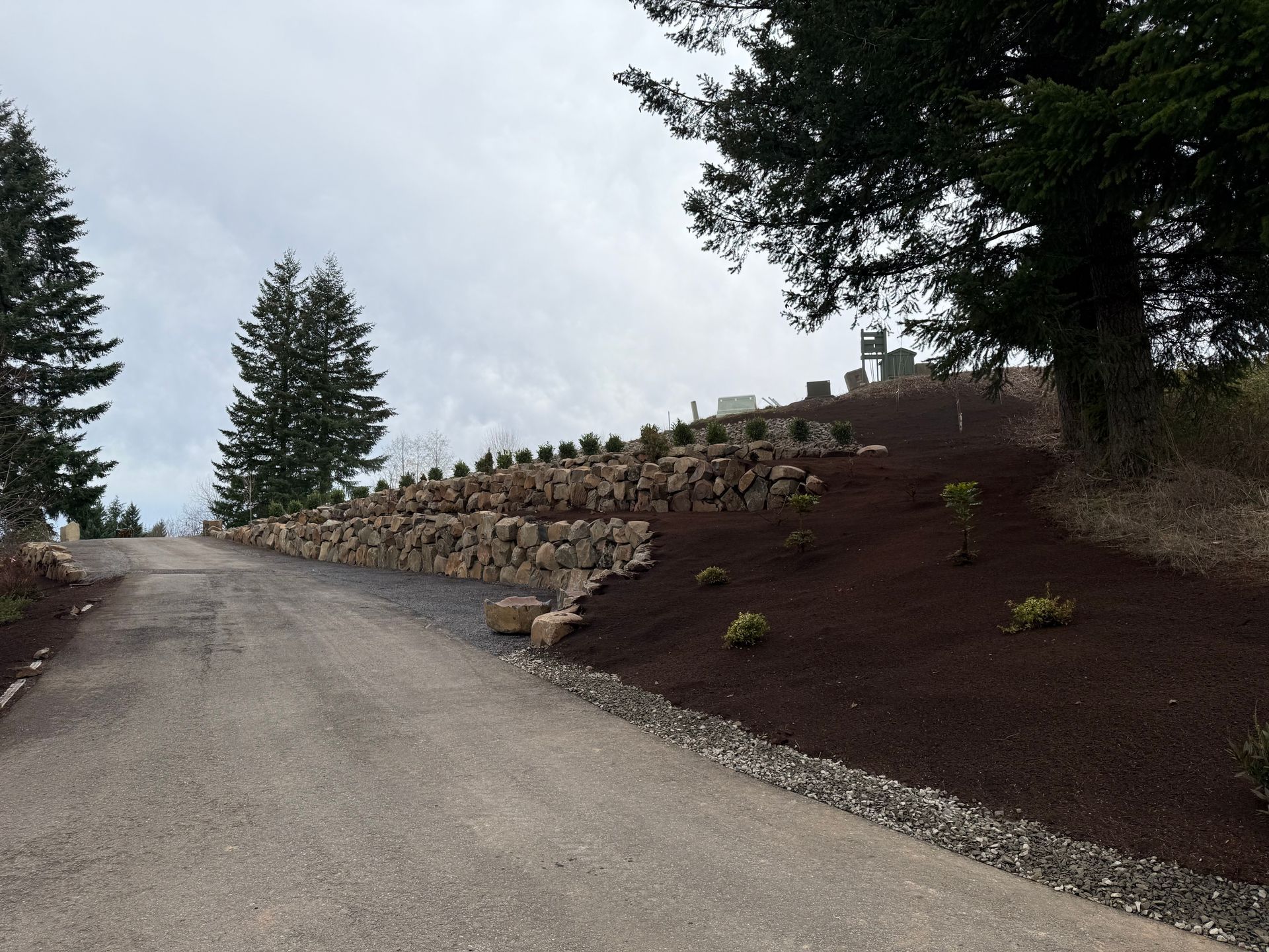 Gravel road ascends a landscaped hillside with stacked stone retaining walls, topped with shrubs, under an overcast sky.