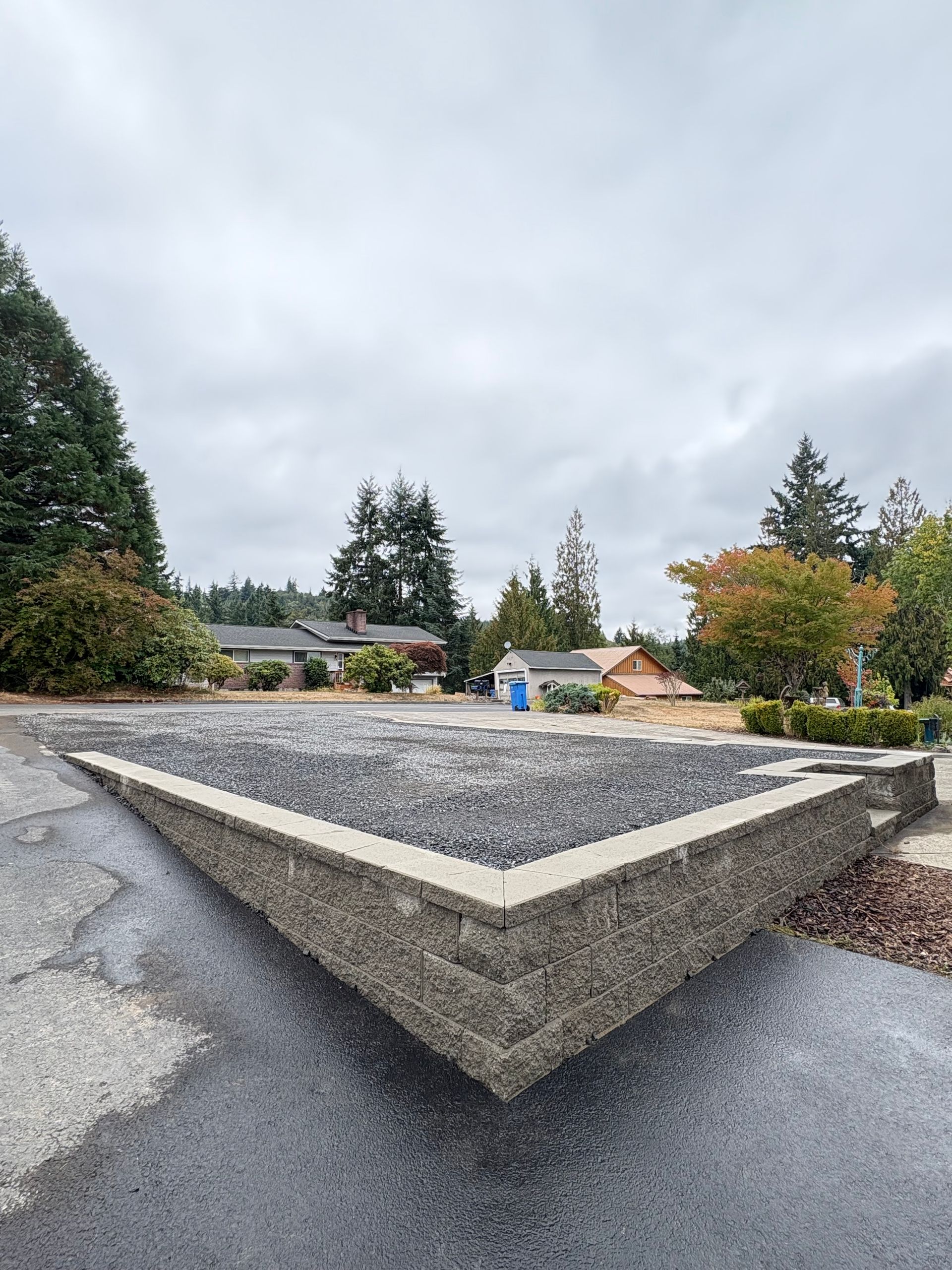 Concrete retaining wall filled with dark gravel; houses and trees in background under overcast sky.