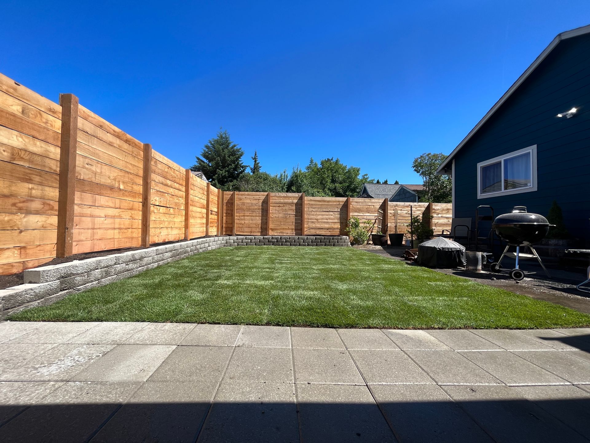 Backyard with green lawn, wooden fence, and blue house under a clear blue sky.