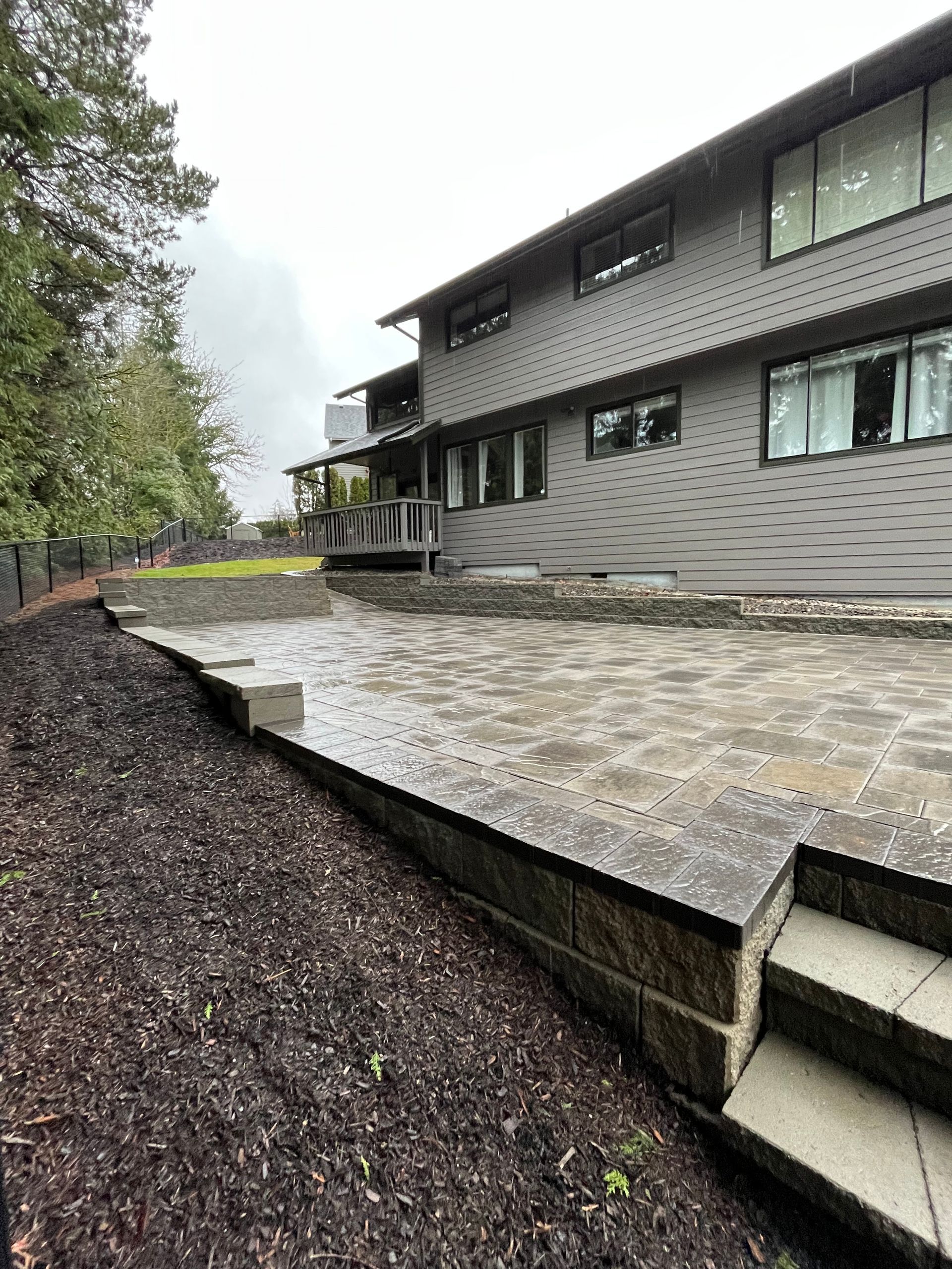 Backyard patio with stone pavers and retaining walls. Two-story gray house in background, steps, and dark mulch.