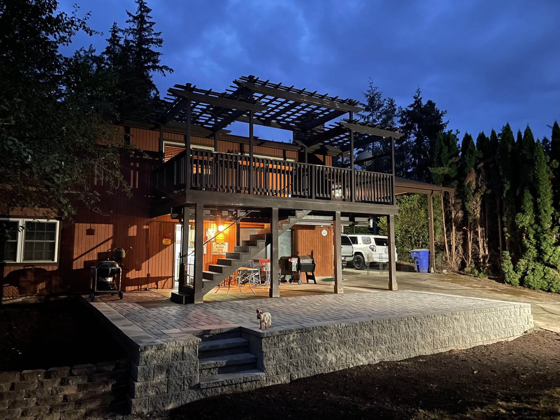 Two-story wooden house with deck and pergola, stone patio, and dark sky.