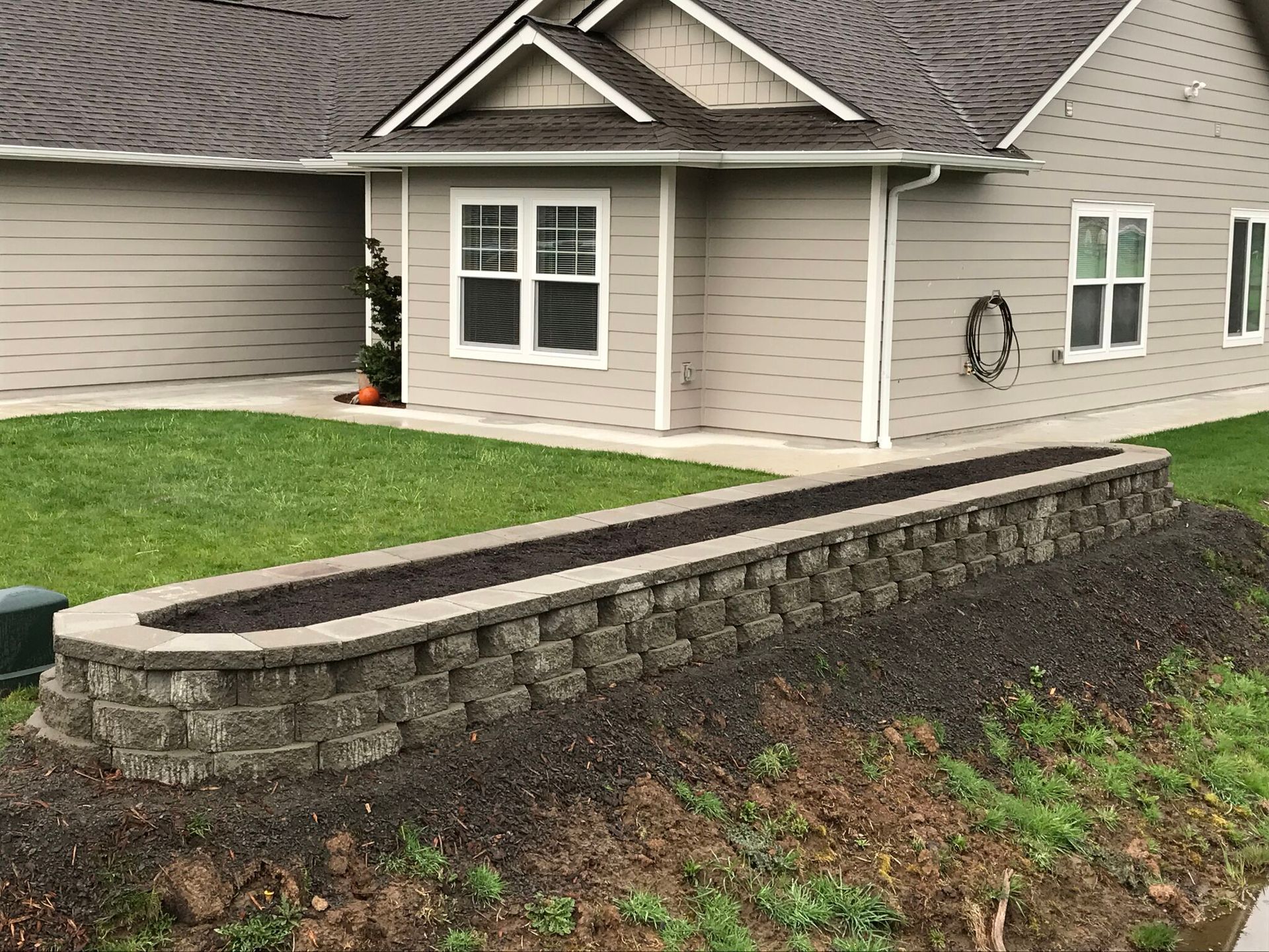 Stone retaining wall with planter filled with dark soil, near a light brown house with green lawn.