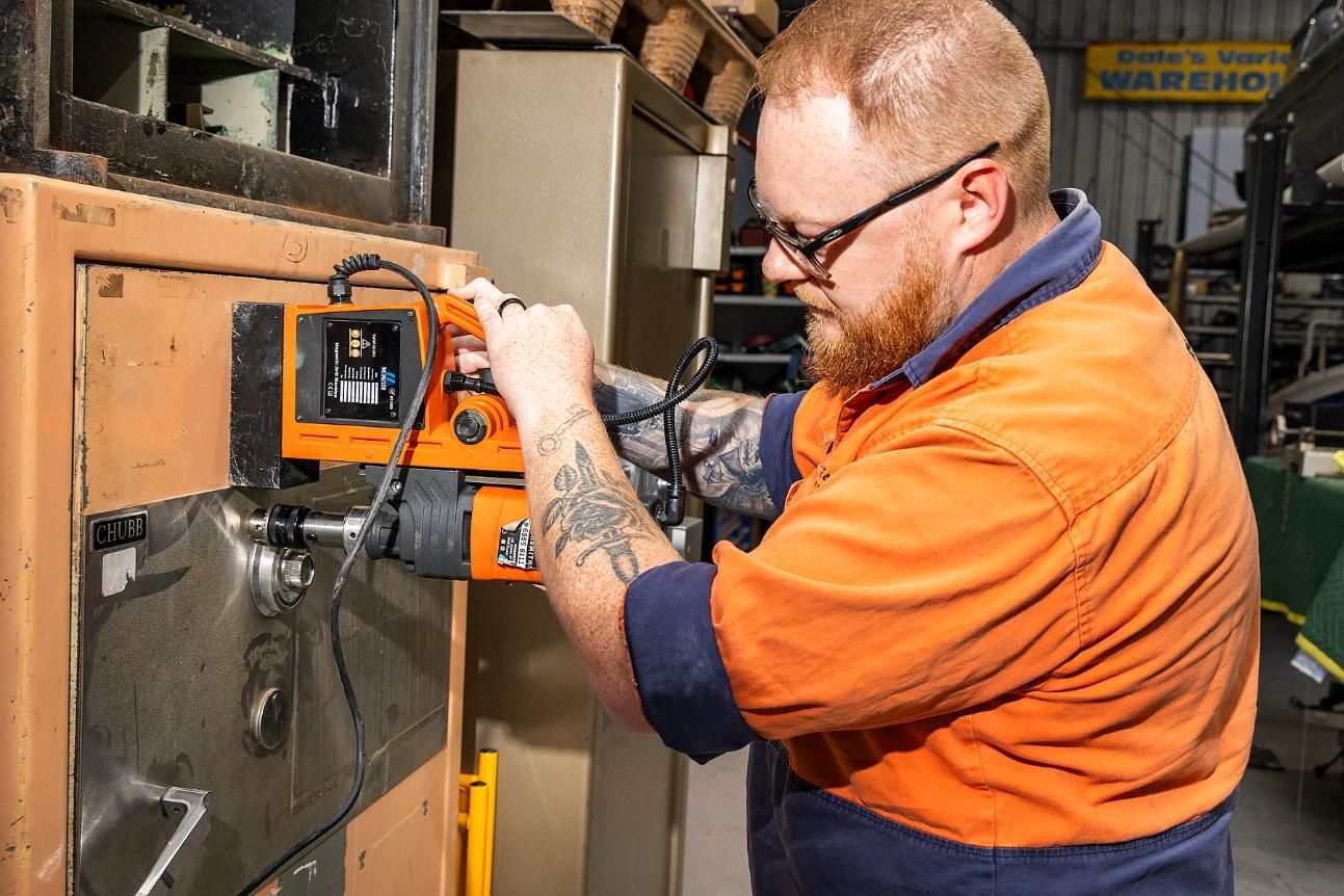 A Man Using Machinery to Open a Safe in a Workshop, Wearing an Orange Shirt and Glasses — Active 24/7 Locksmith and Security Services in Forster, NSW