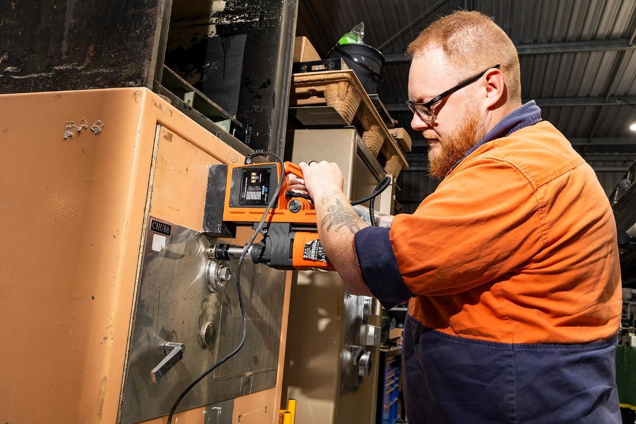 Man in Orange Work Shirt Drills Into Metal in a Workshop — Active 24/7 Locksmith and Security Services in Forster, NSW