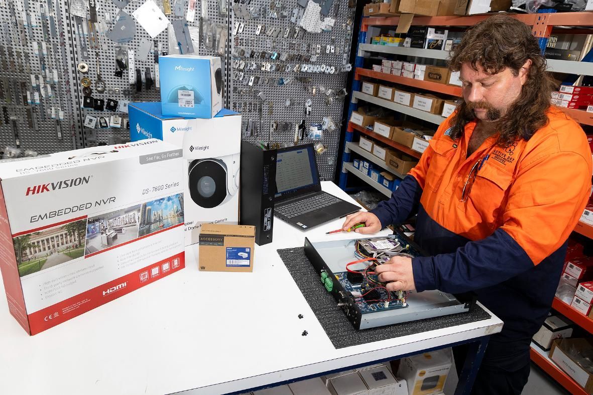 Man With Mullet Working on Electronics at a Table, Surrounded by Boxes and Shelves in a Warehouse Setting — Active 24/7 Locksmith and Security Services in Forster, NSW