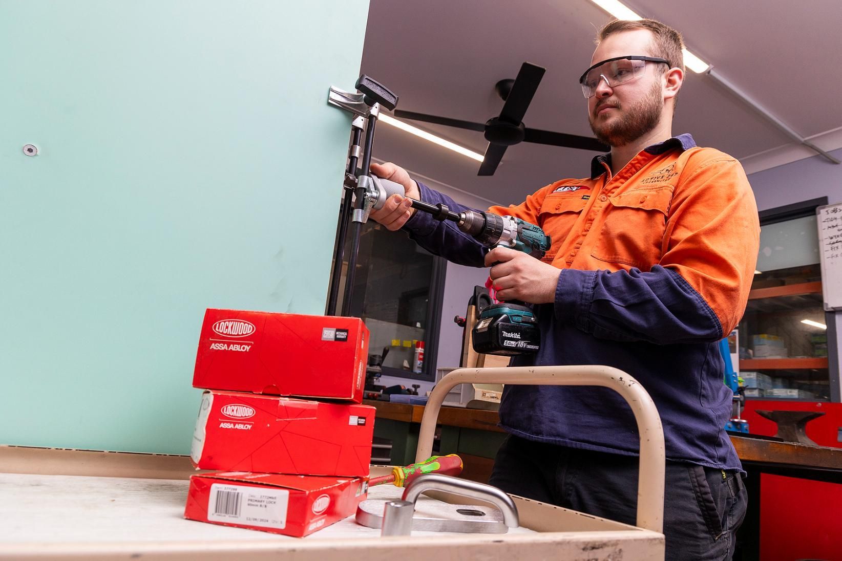Man in Safety Glasses Uses a Drill on Wall, Boxes of Supplies on a Cart — Active 24/7 Locksmith and Security Services in Bulahdelah, NSW