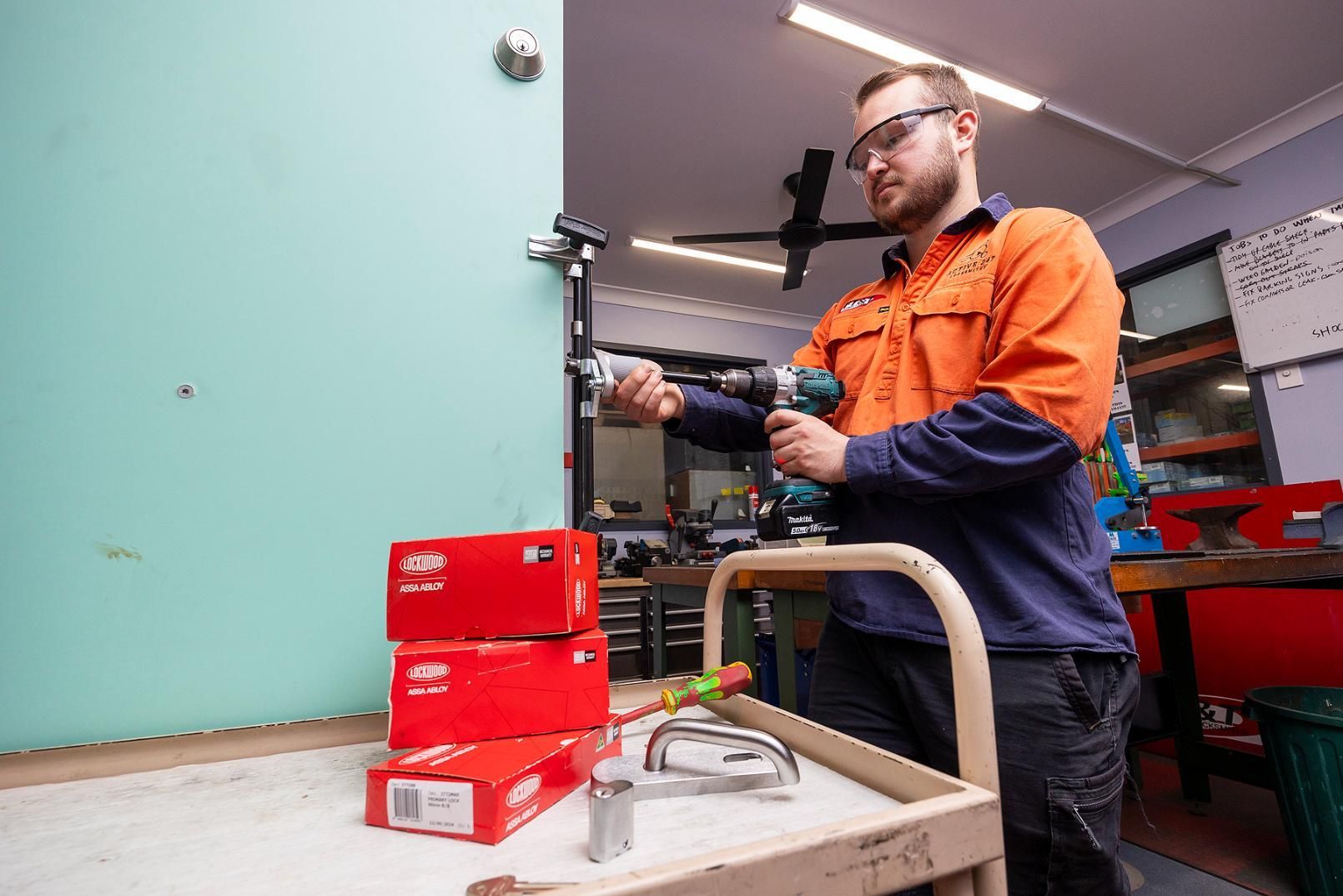 Man in Work Clothes Drilling a Door. Boxes of Parts Sit on a Cart in a Workshop — Active 24/7 Locksmith and Security Services in Nabiac, NSW