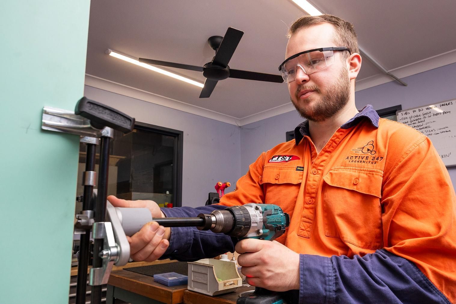 Man in Safety Glasses Drilling Into a Door Frame, Orange Shirt, in a Workshop — Active 24/7 Locksmith and Security Services in Gloucester, NSW
