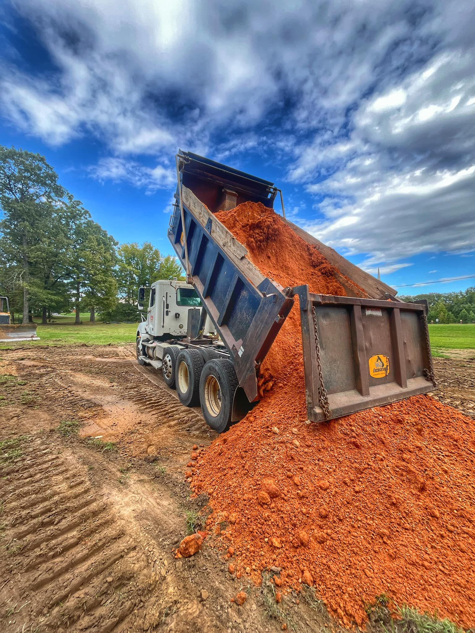 A dump truck with its bed raised, emptying a large pile of red mulch onto a dirt construction site under a cloudy sky.
