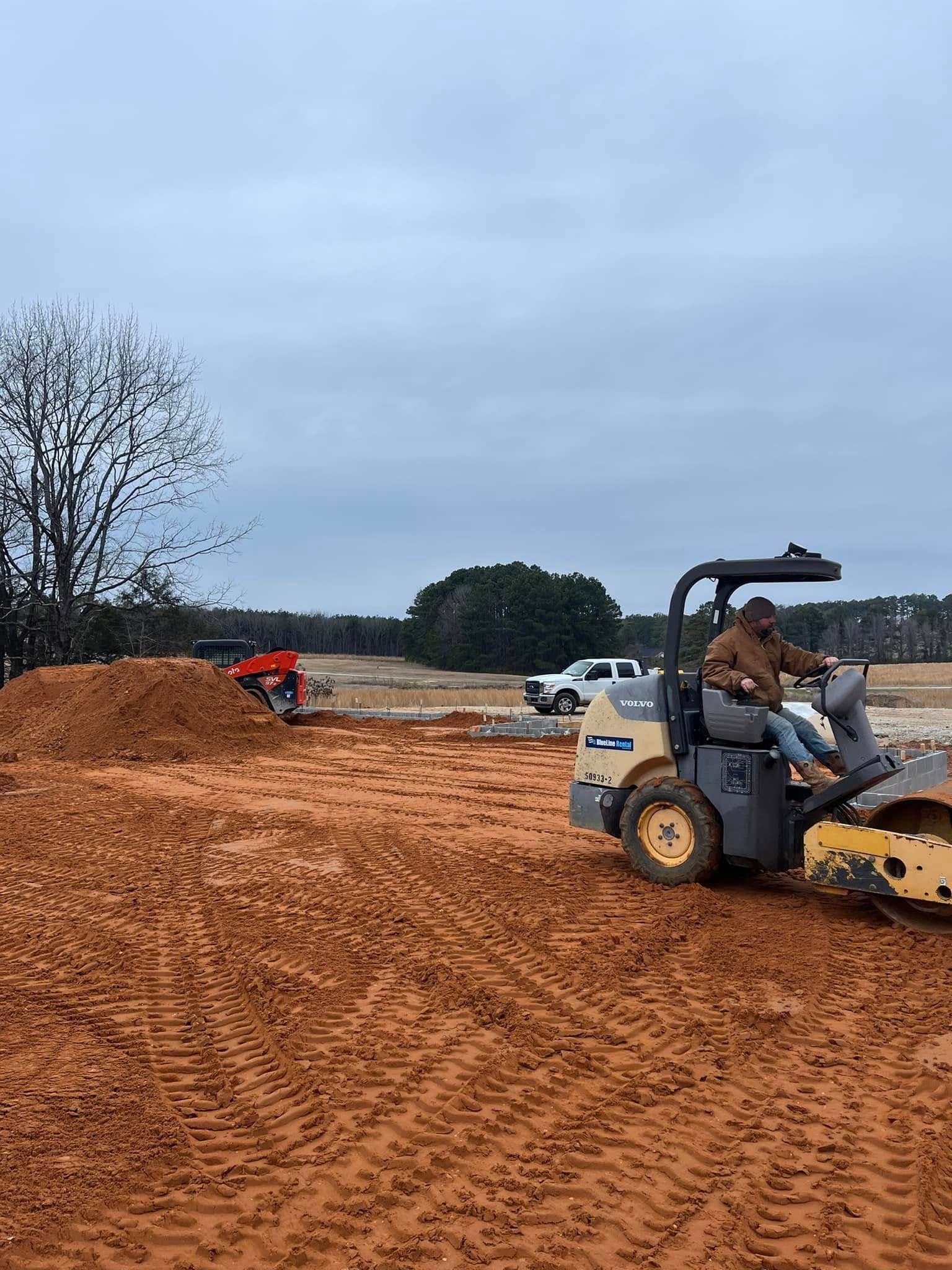 A person operates a yellow soil compactor on a large dirt construction site under an overcast sky.