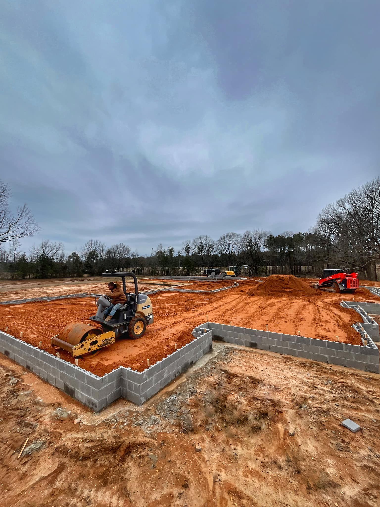 A construction worker operates a yellow soil compactor inside the block foundation of a building site with red dirt.