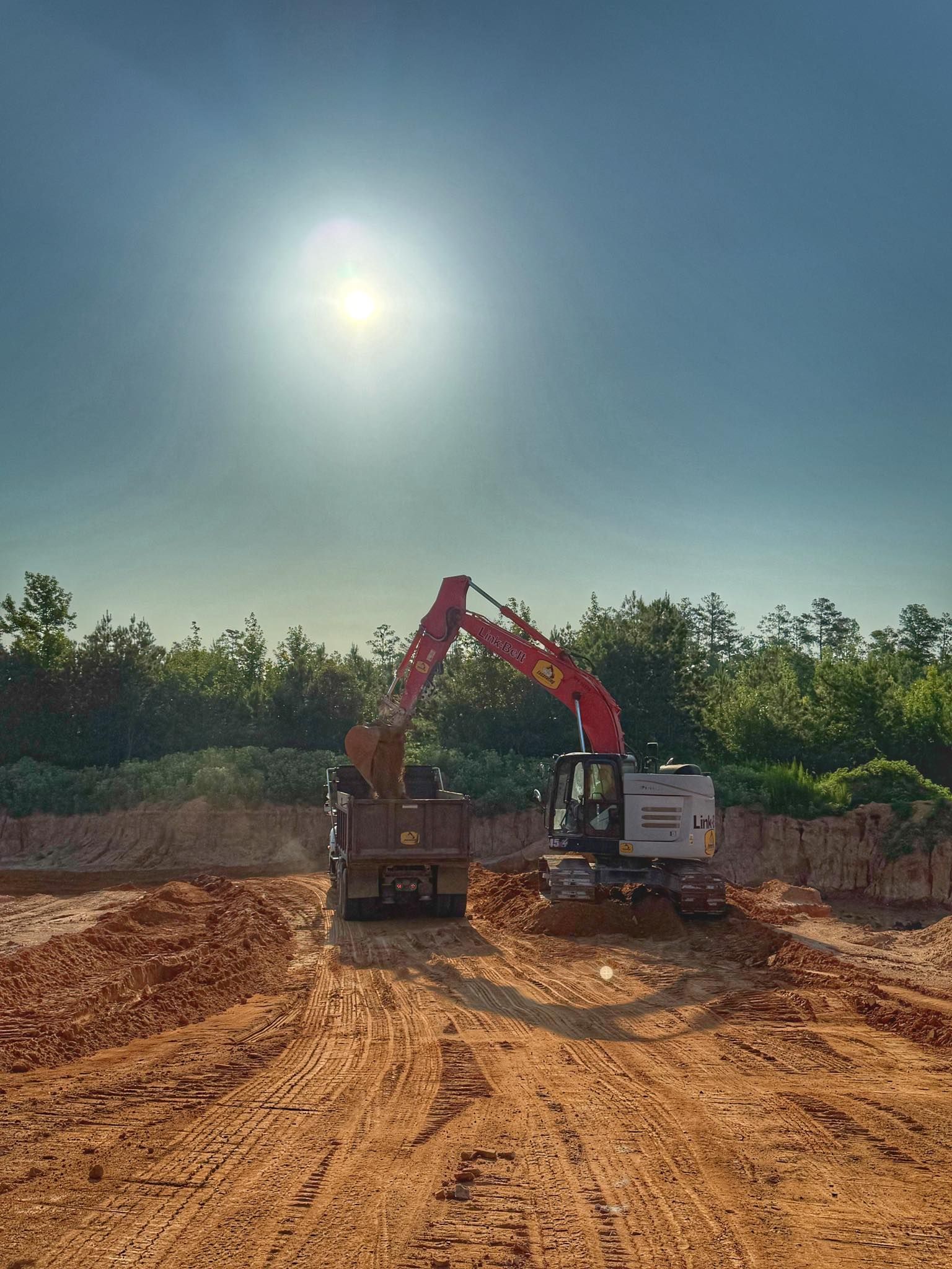 A red excavator loads dirt into a dump truck on a sunny, dirt-filled construction site surrounded by trees.